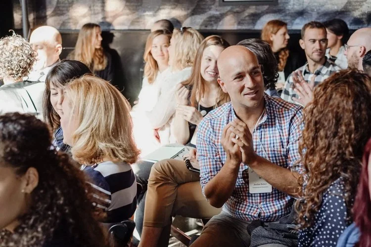 Group of diverse people attending a conference or seminar, seated in a dark room, listening, clapping, and smiling.