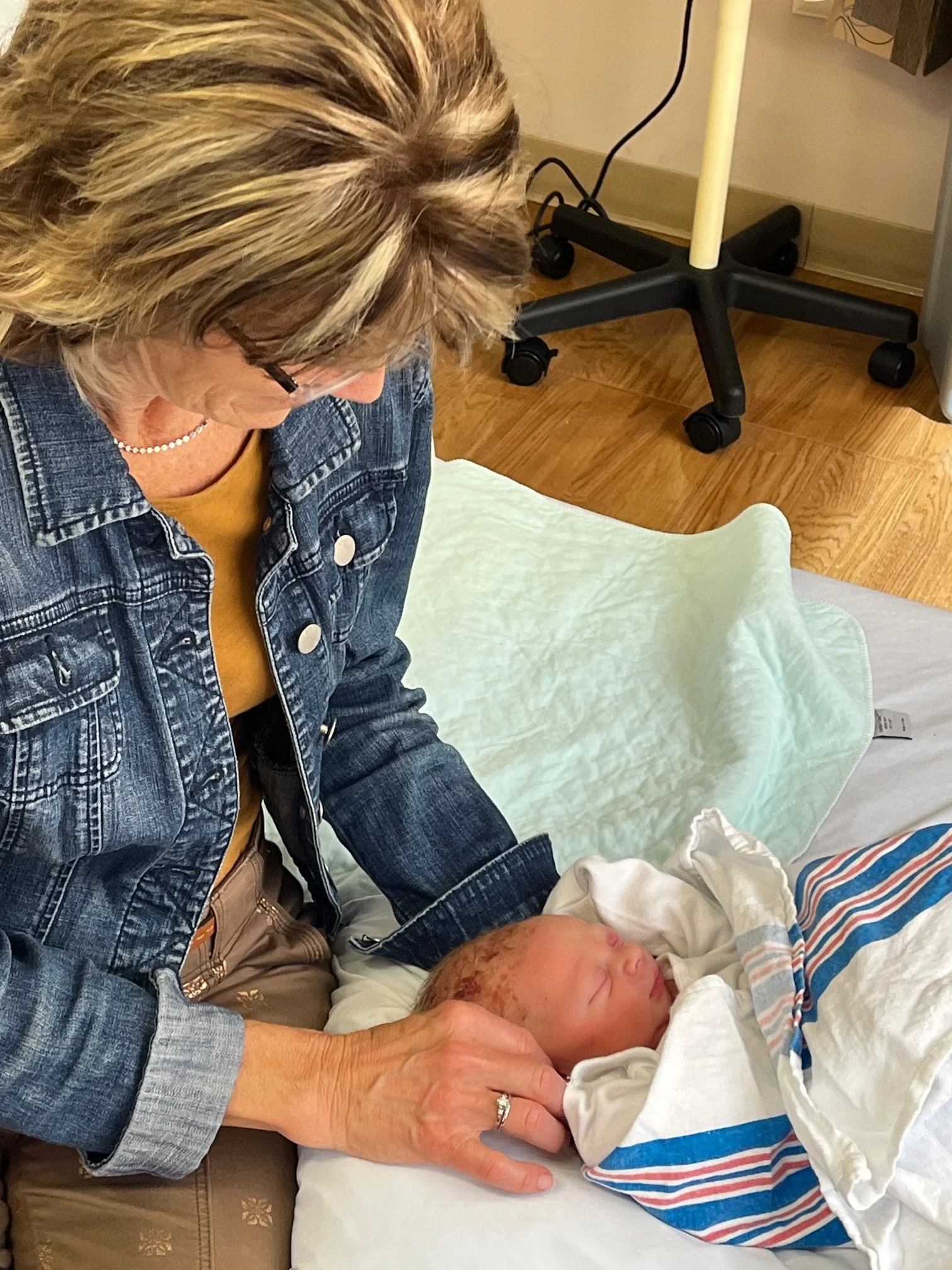 An older woman with short, blonde, and gray hair wearing glasses and a denim jacket holds a newborn baby wrapped in a hospital blanket on a hospital bed. The woman is looking down at the baby in a nurturing manner.