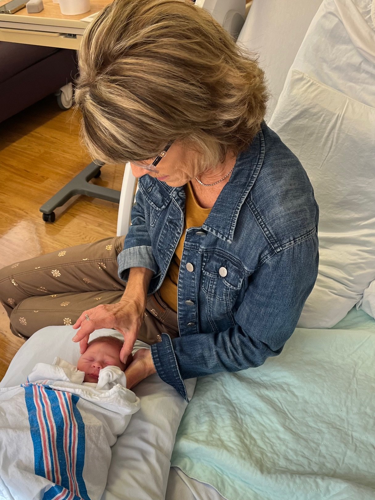 An older woman with glasses and a denim jacket gently touches the forehead of a newborn baby wrapped in a hospital blanket, sitting on a hospital bed.