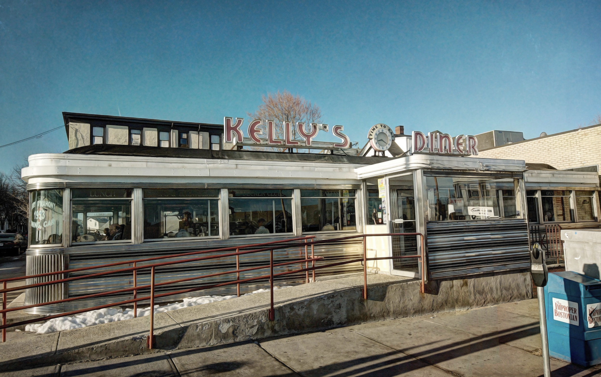 Exterior of Kelly's Diner with vintage design, large windows, and neon sign on roof, on a clear day with blue sky.