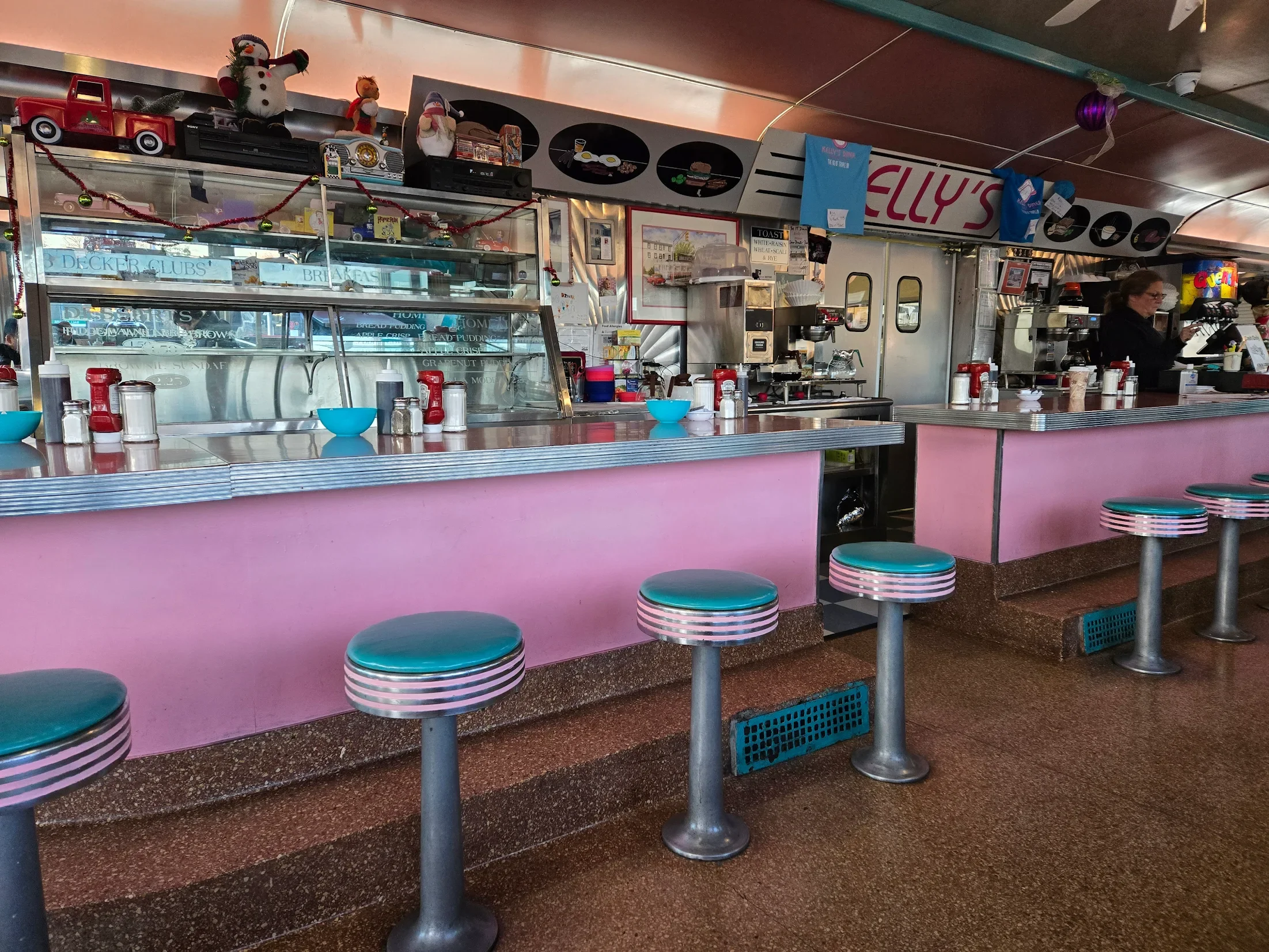 Inside of Kelly's Diner, with pink counter, turquoise stools, and retro decor. There are napkin dispensers on the counter, Christmas decorations, and a woman working at the counter.