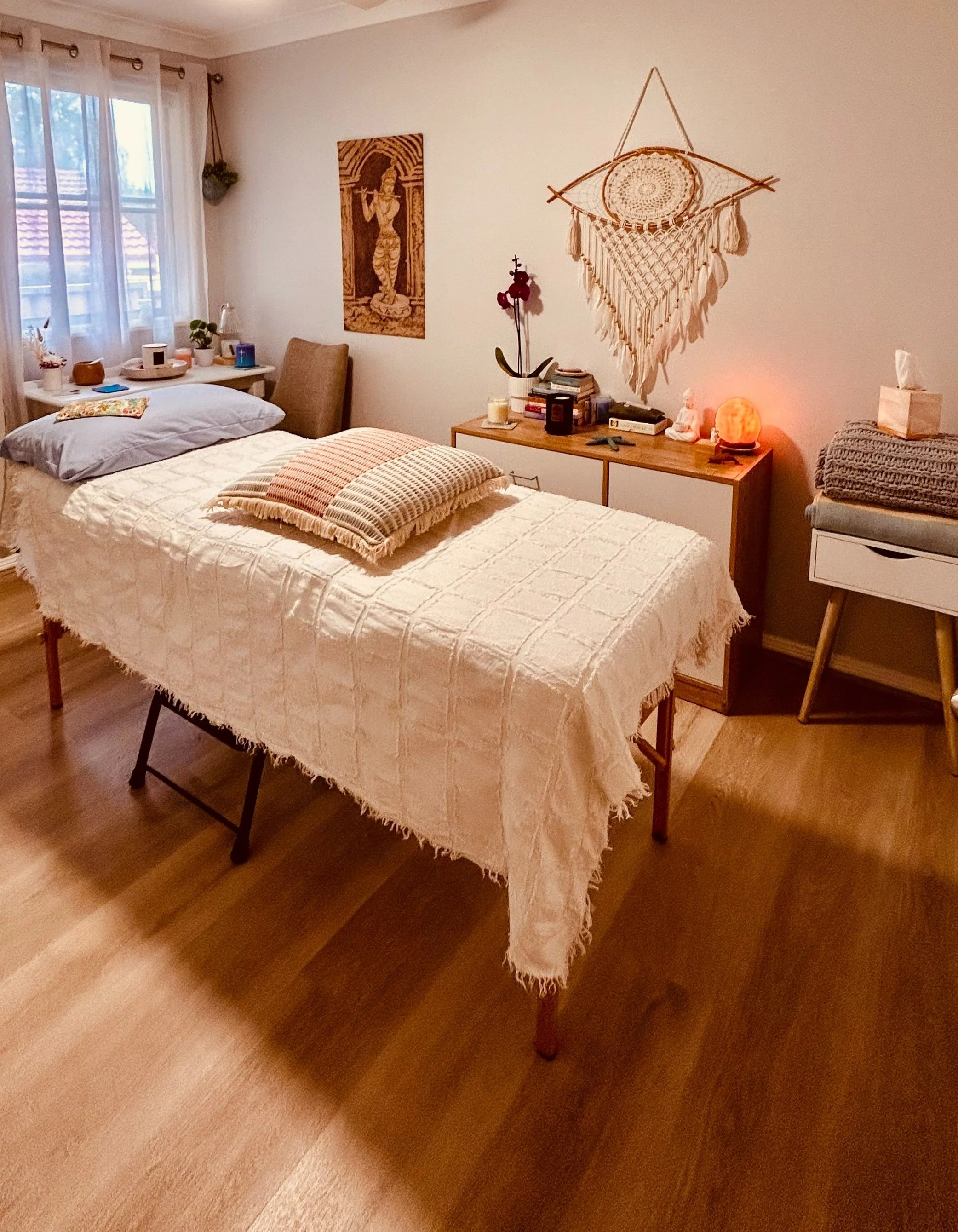 A cozy bedroom with a bed covered in a cream-colored blanket, and pillows on top, near a window with sheer curtains. There's a nightstand with plants, decor, and books, and a wall hanging with a dream catcher design. A salt lamp is also on the nightstand. A small table on the right holds neatly folded blankets and tissues.