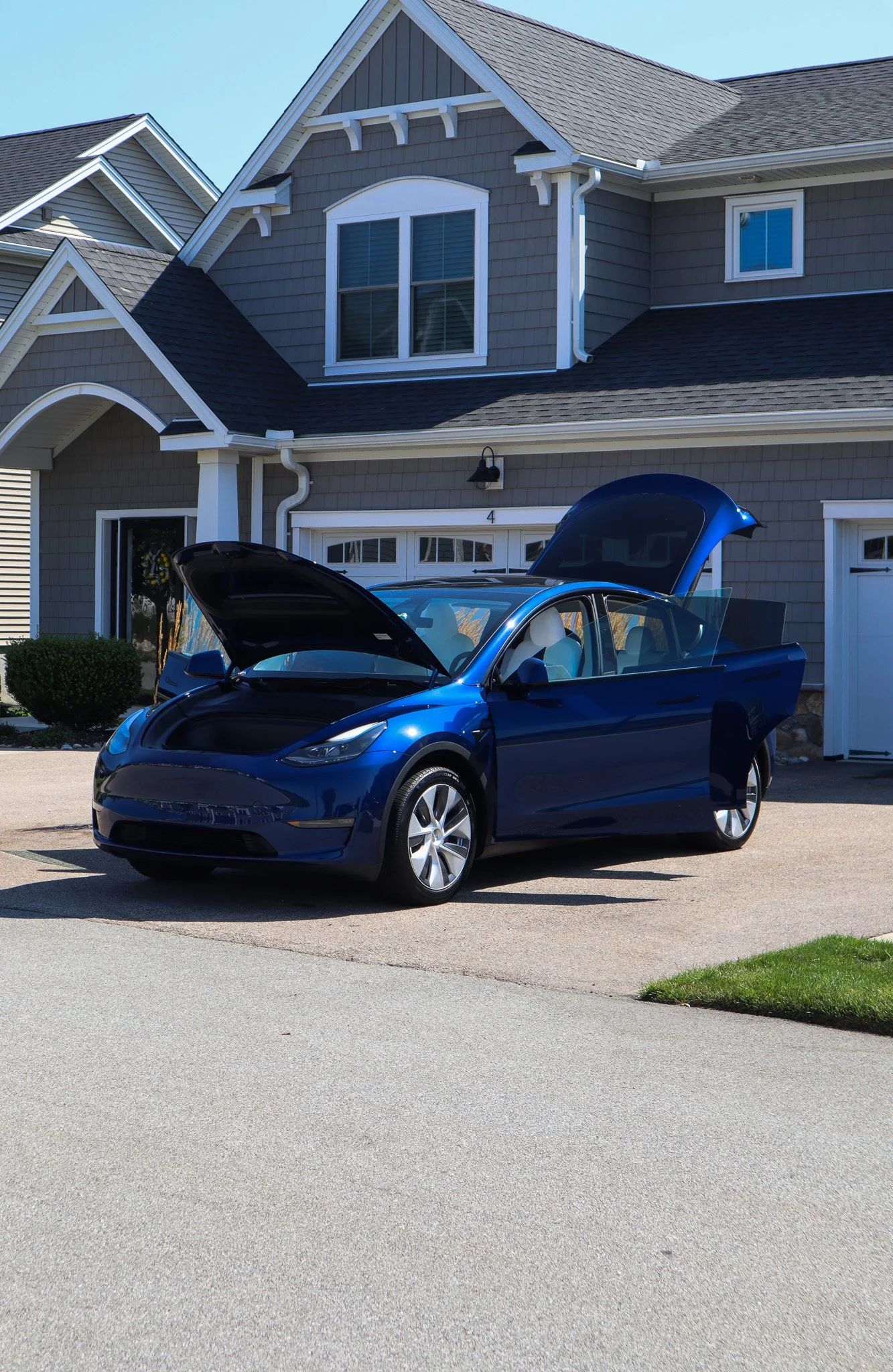 A blue Tesla Model 3 parked in a driveway in front of a gray house with open hood and trunk.