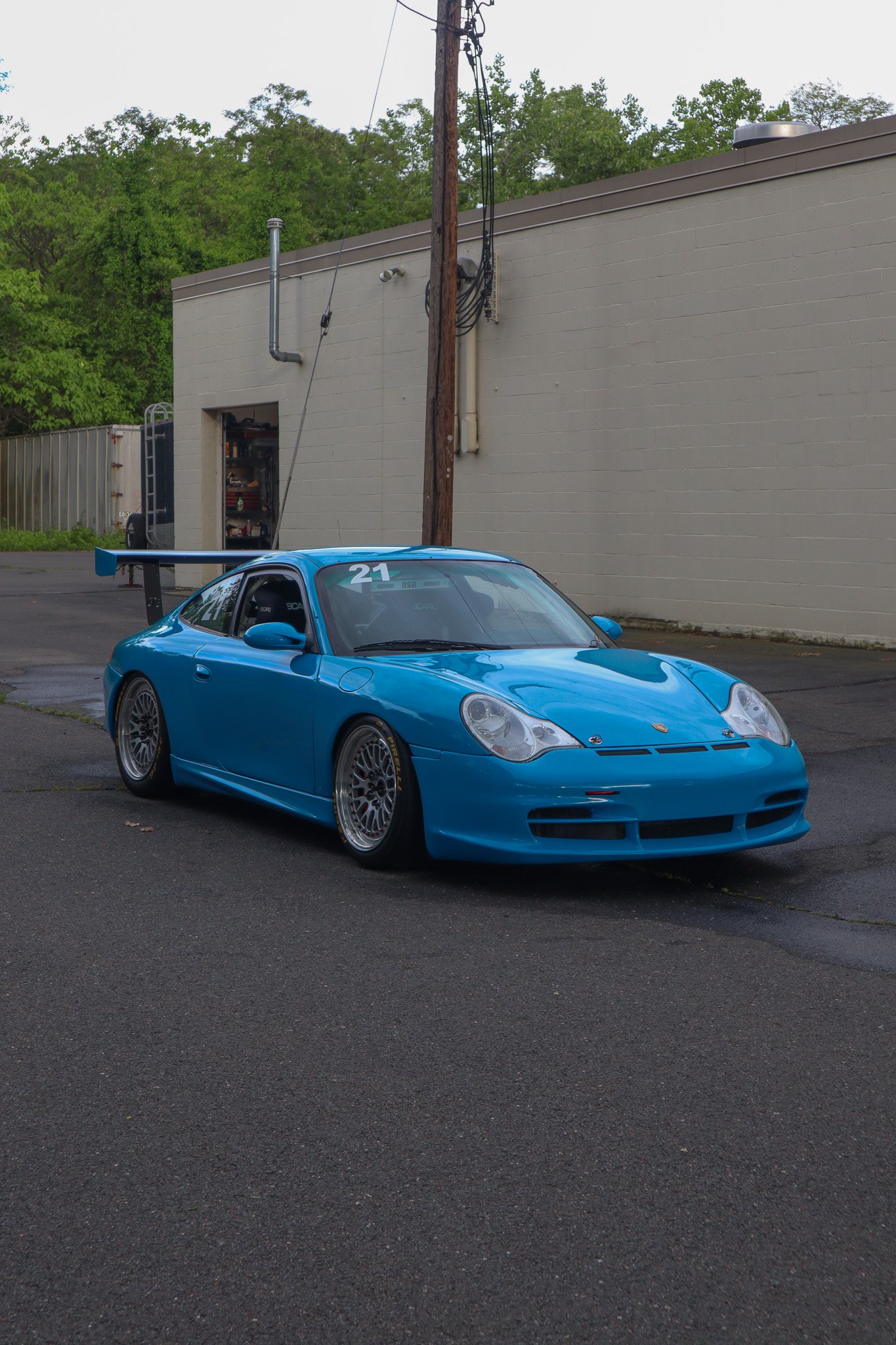 A blue Porsche race car with the number 21 on the windshield parked on an asphalt surface near a beige building with green trees in the background.