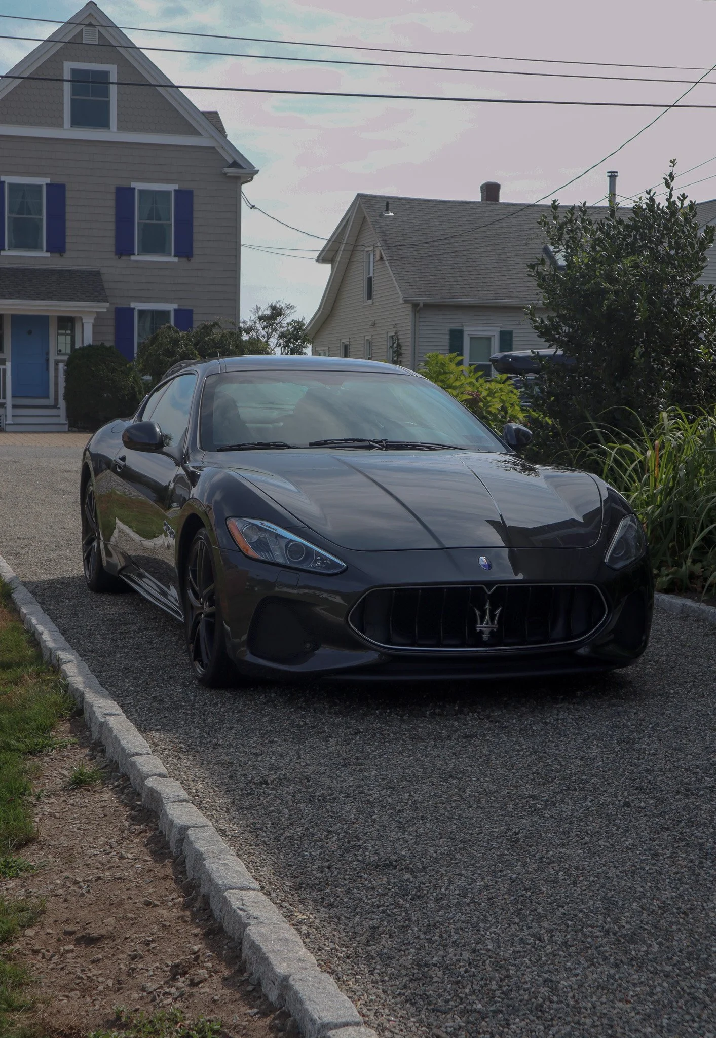 A black Maserati sports car parked in a residential driveway with houses and bushes in the background.