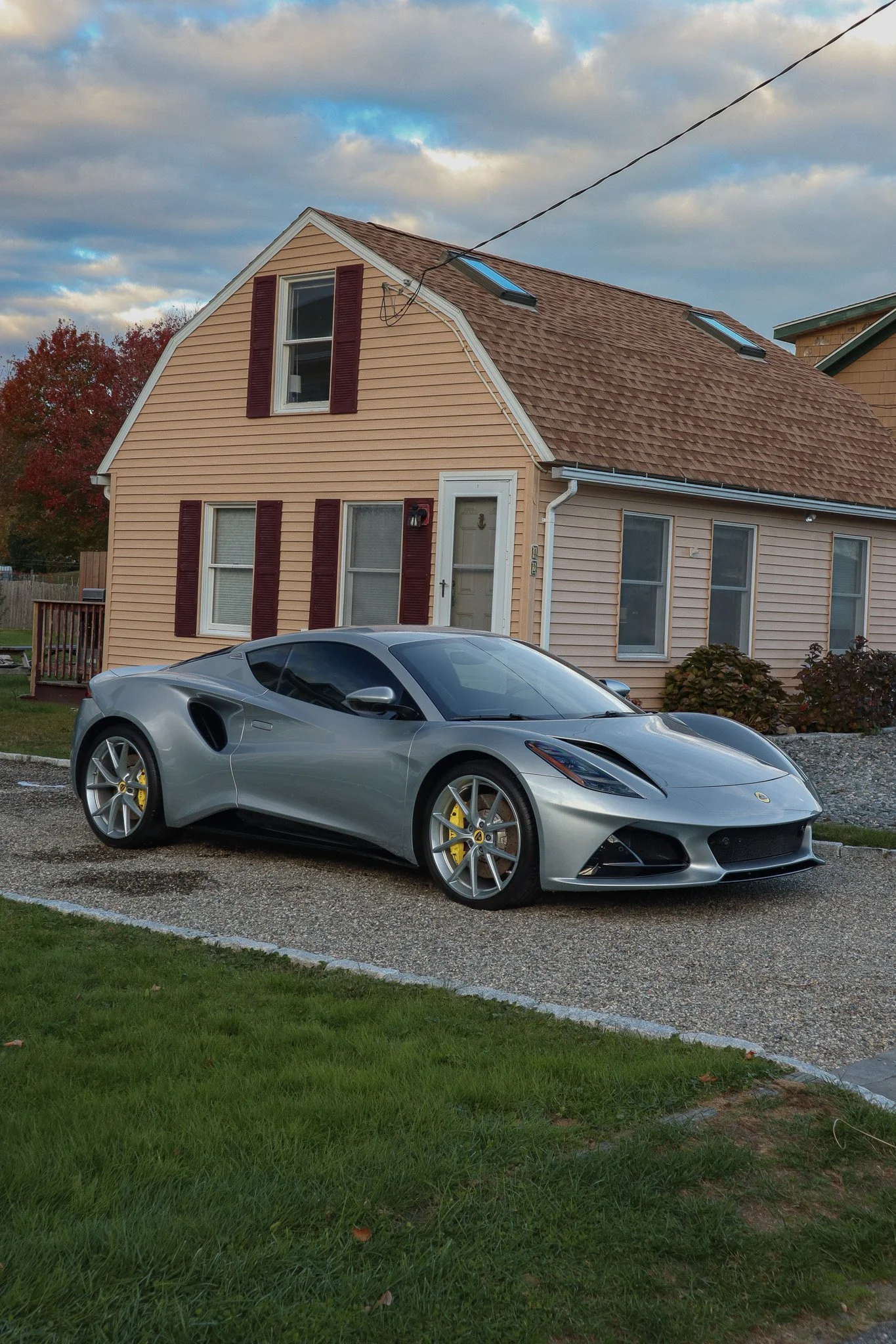 A silver sports car parked on a gravel driveway in front of a peach-colored house with maroon shutters and a gabled roof, with clouds in the sky.
