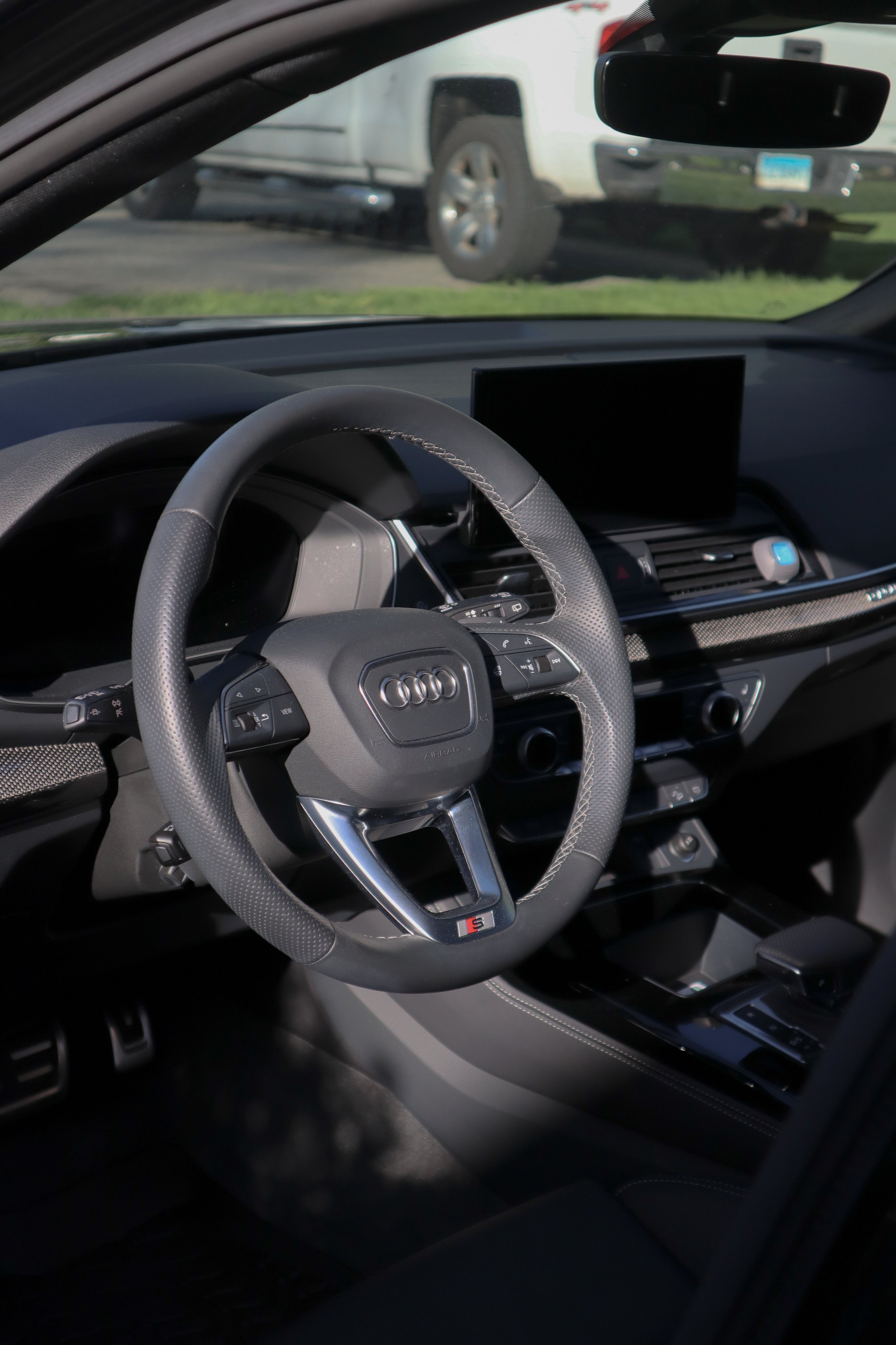 Interior of an Audi vehicle, focusing on the steering wheel and dashboard with a large screen display, with a white vehicle visible outside through the windshield.