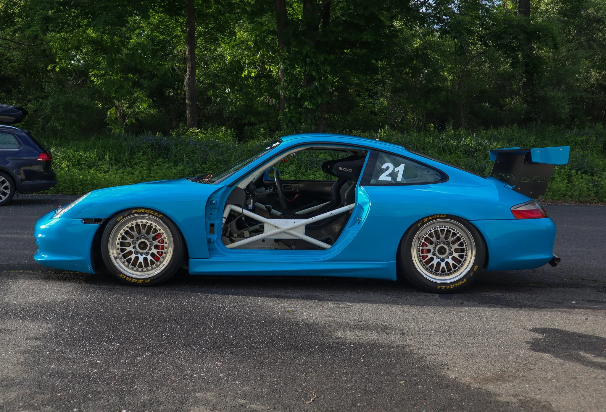 A blue Porsche 911 GT3 cup car parked on the side of the road with the driver's side door removed, exposing the interior and racing roll cage, in front of a background of green trees. Showcasing the elegance of Doorstep Detailing's astonishing work.