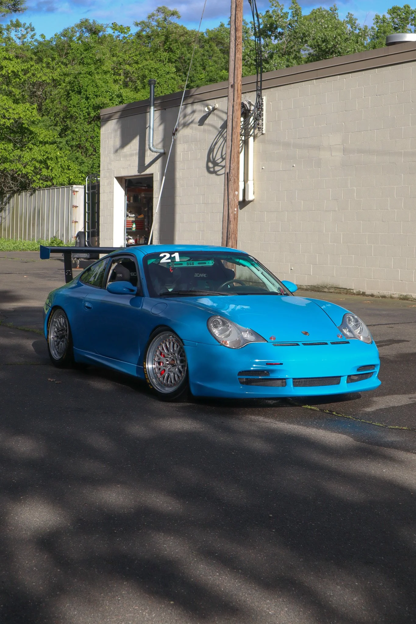 A blue Porsche race car with the number 21 on the windshield, parked outside near a beige building with a garage door open, and a sign that says BSR inside the car.