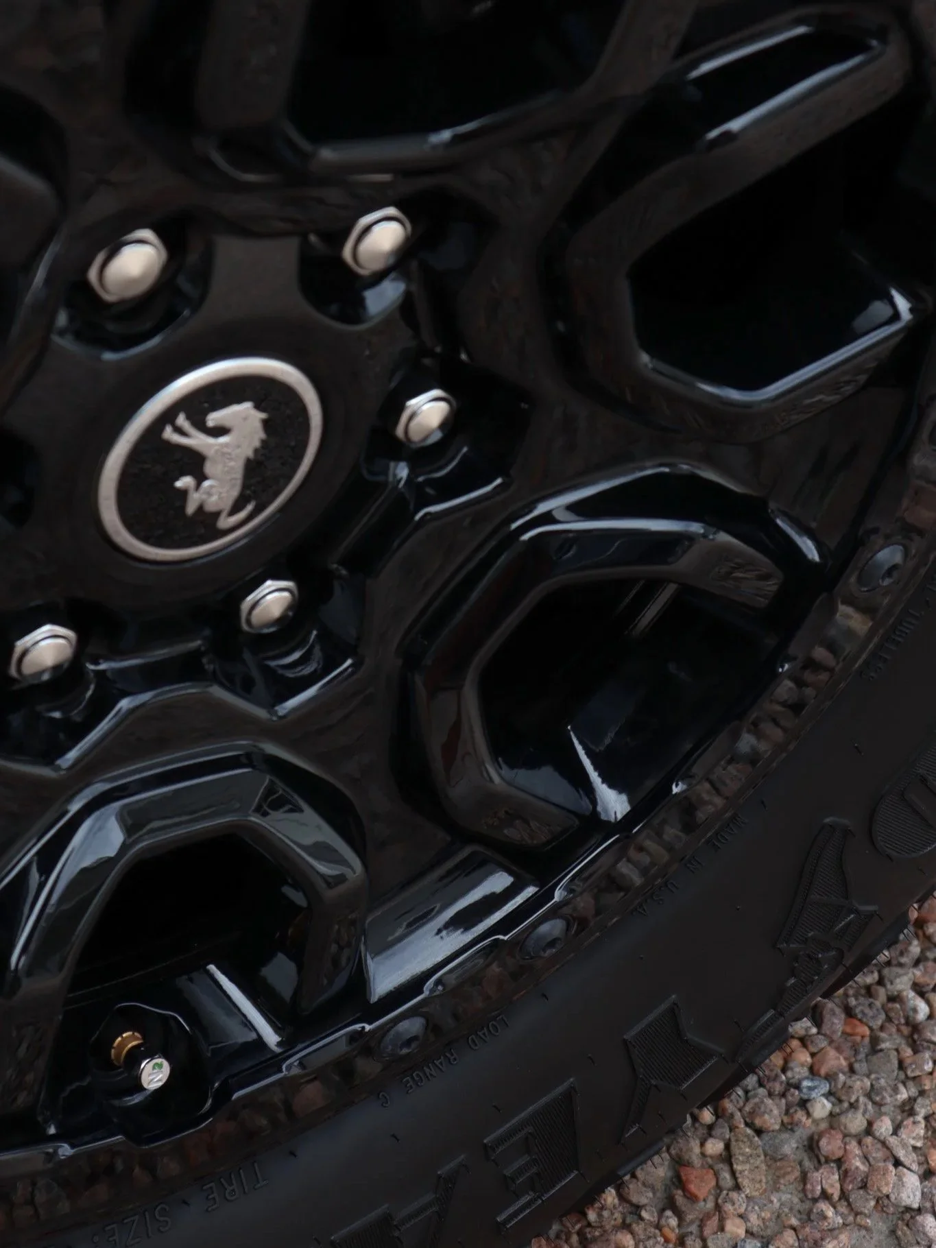 Close-up of a black car wheel with a logo of a horse rearing on the hubcap, and the tire tread visible on the gravel surface.