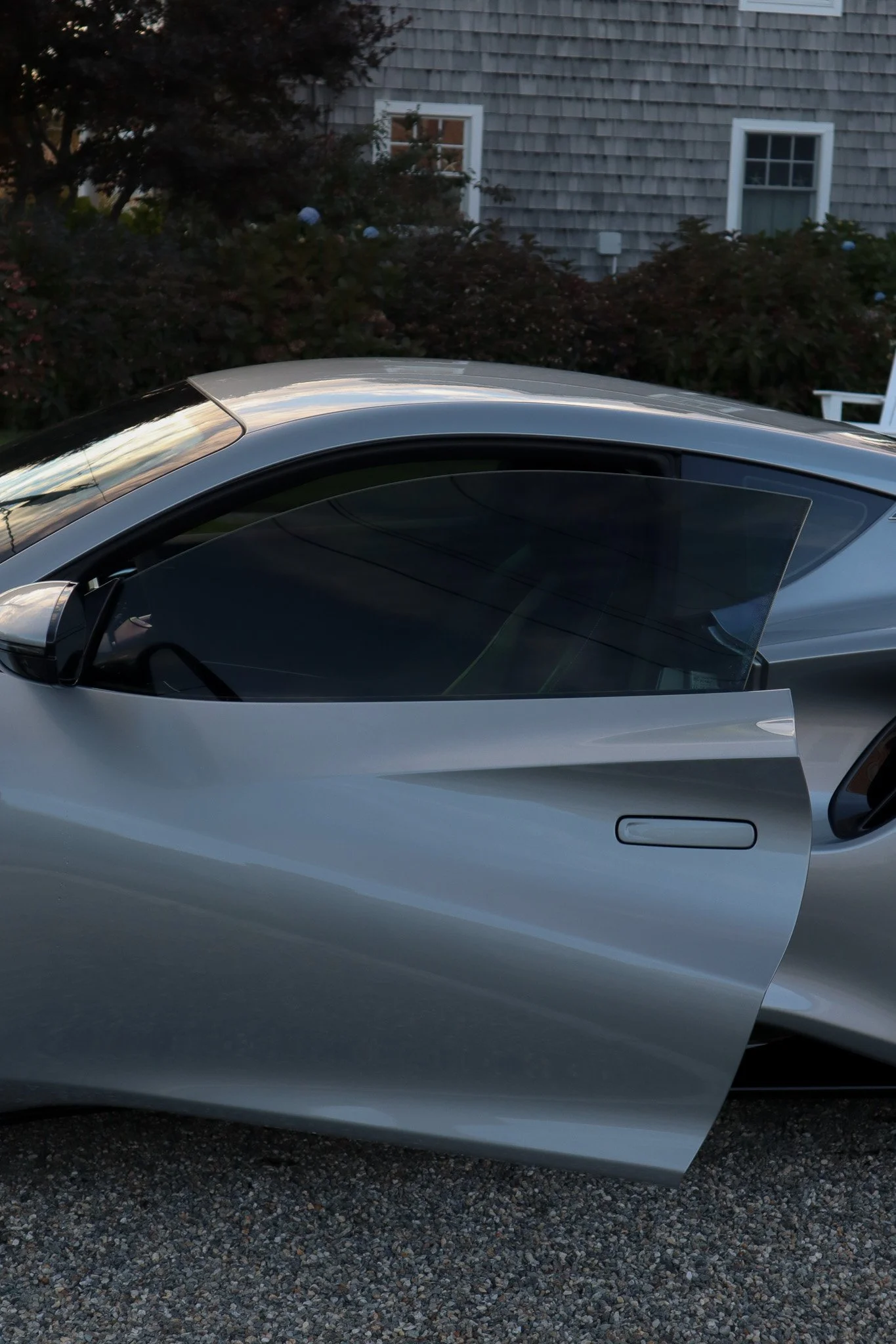 A silver sports car with a racing-style door open, parked on a driveway with a house and bushes in the background.