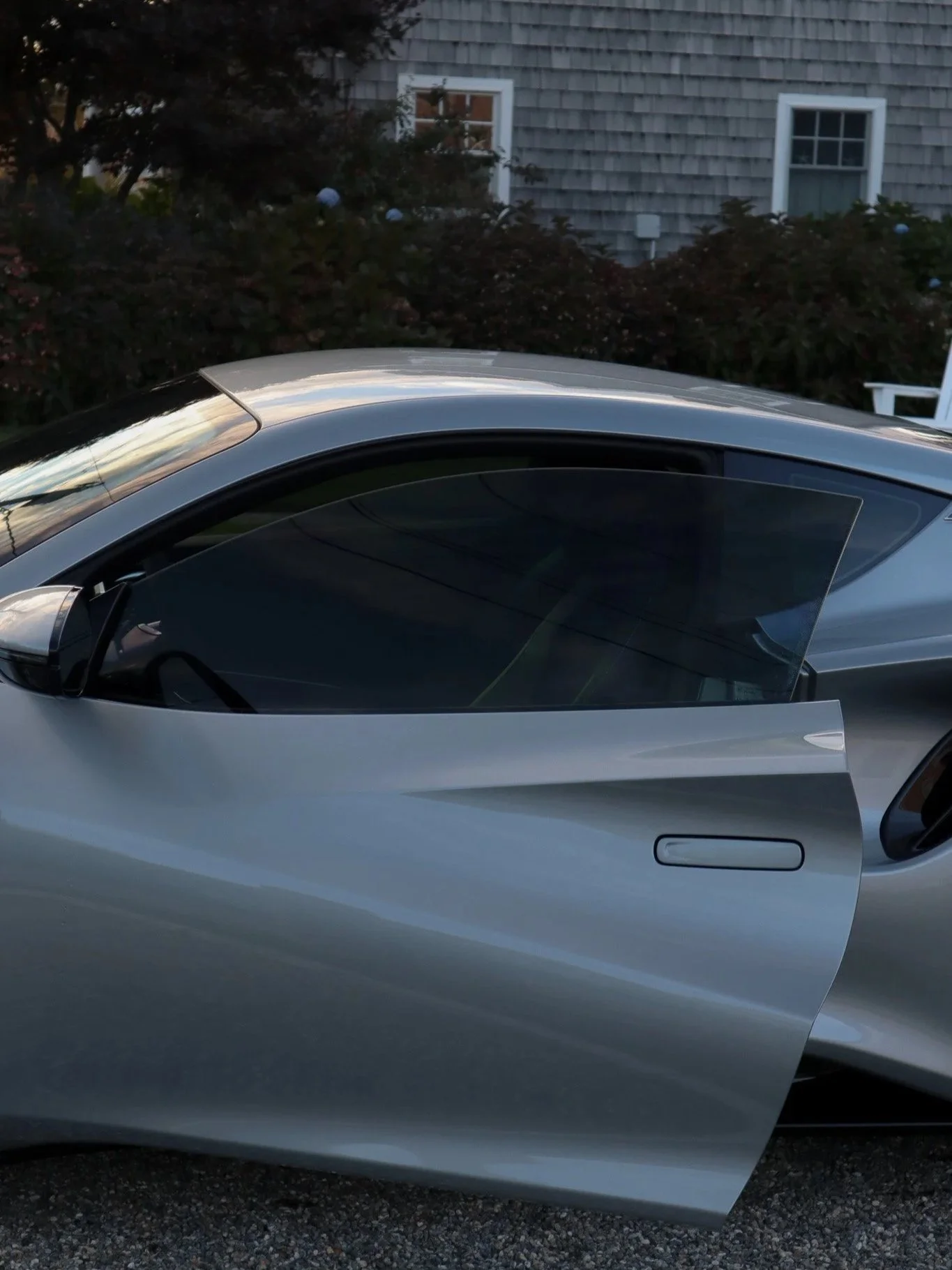 Silver sports car with an open driver's side door parked in front of a house with gray shingles and white-framed windows, surrounded by bushes and trees.