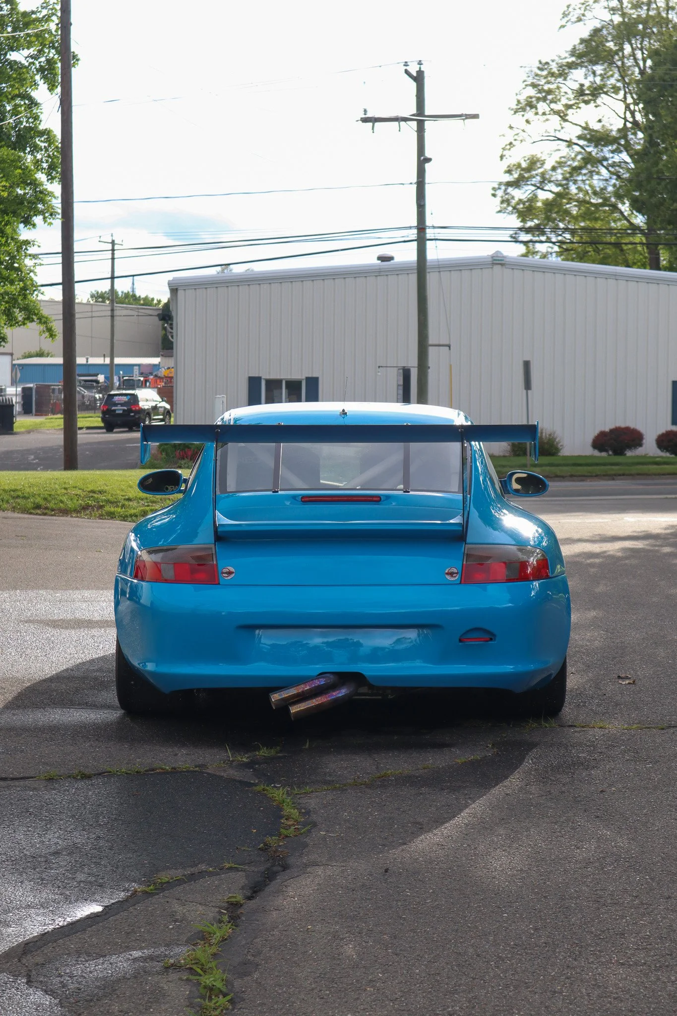 rear view of a bright blue sports car with a large rear wing and dual exhaust pipes, parked on a cracked asphalt surface with some grass growing through the cracks, in a parking lot with some greenery and industrial buildings in the background.