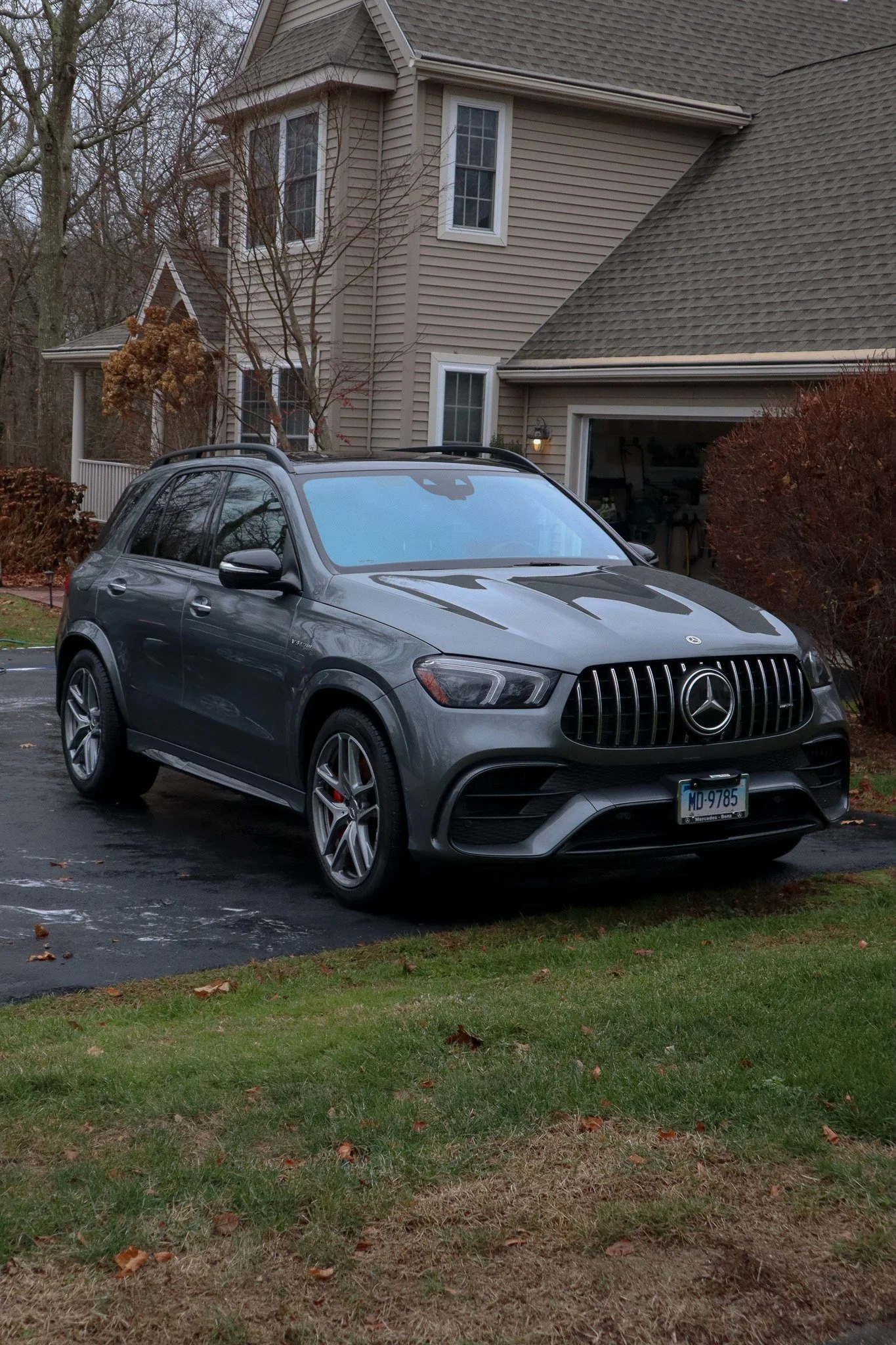 A gray Mercedes-Benz SUV parked on a driveway in front of a beige house with a garage, surrounded by trees with fall foliage, during overcast weather.