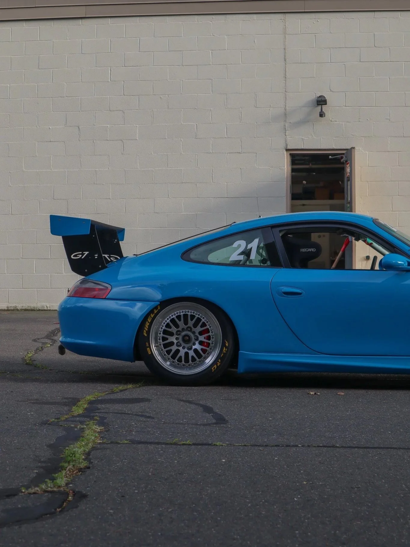 Side view of a blue race car with a rear spoiler, number 21 on the window, parked against a beige brick wall.