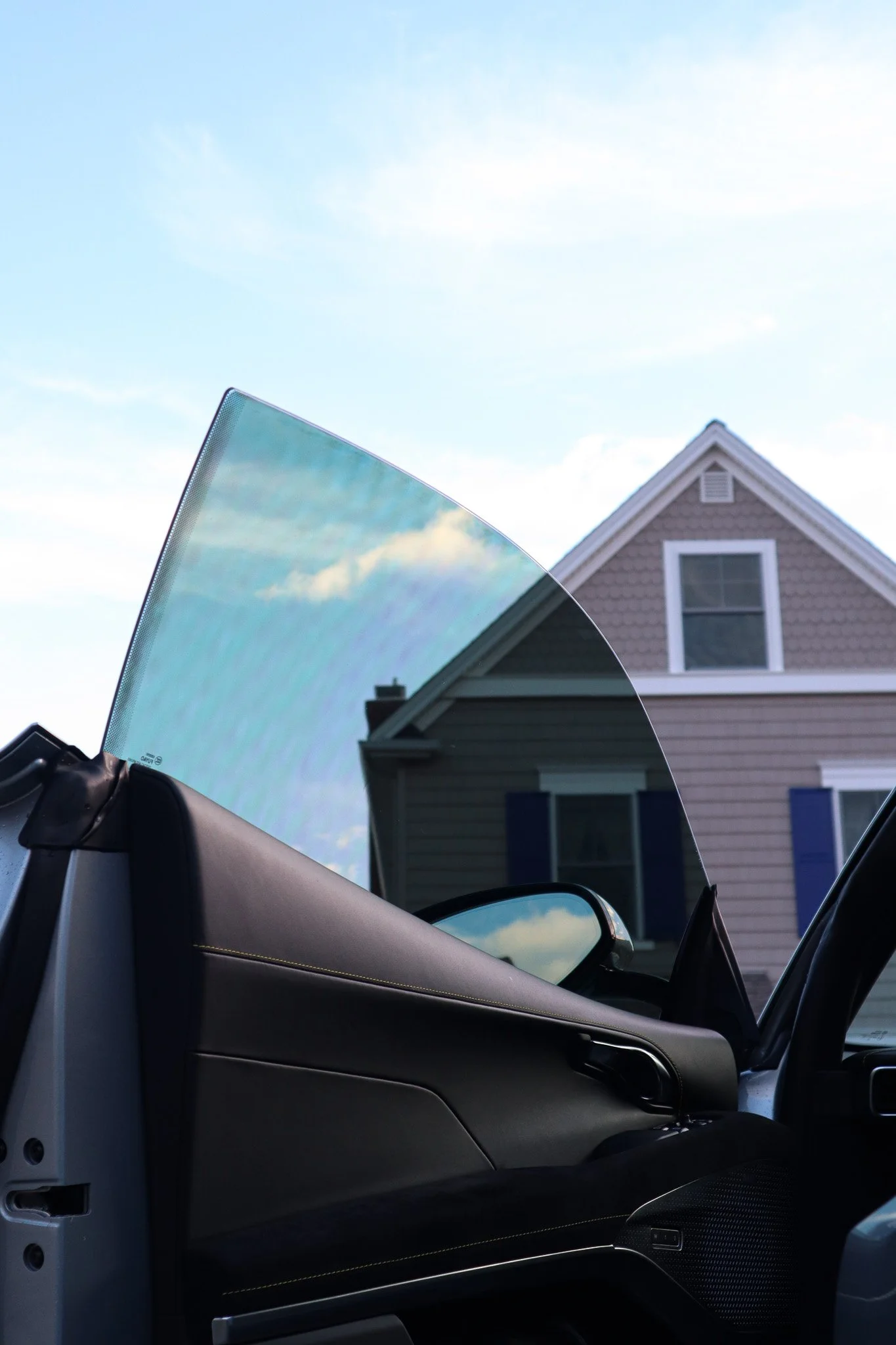 Inside view of a convertible car with the top down, showing the dashboard, with a house with blue shutters and a peaked roof in the background, and blue sky with clouds.