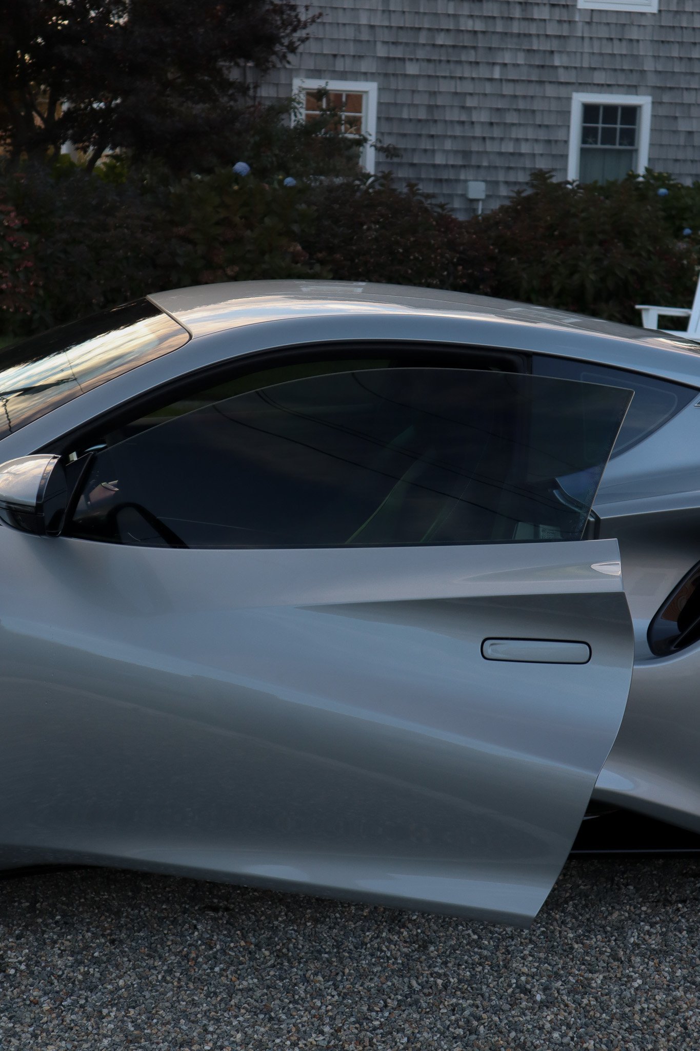 A silver sports car with a partially open door parked outside near a house with a wooden shingle exterior and bushes in the background.