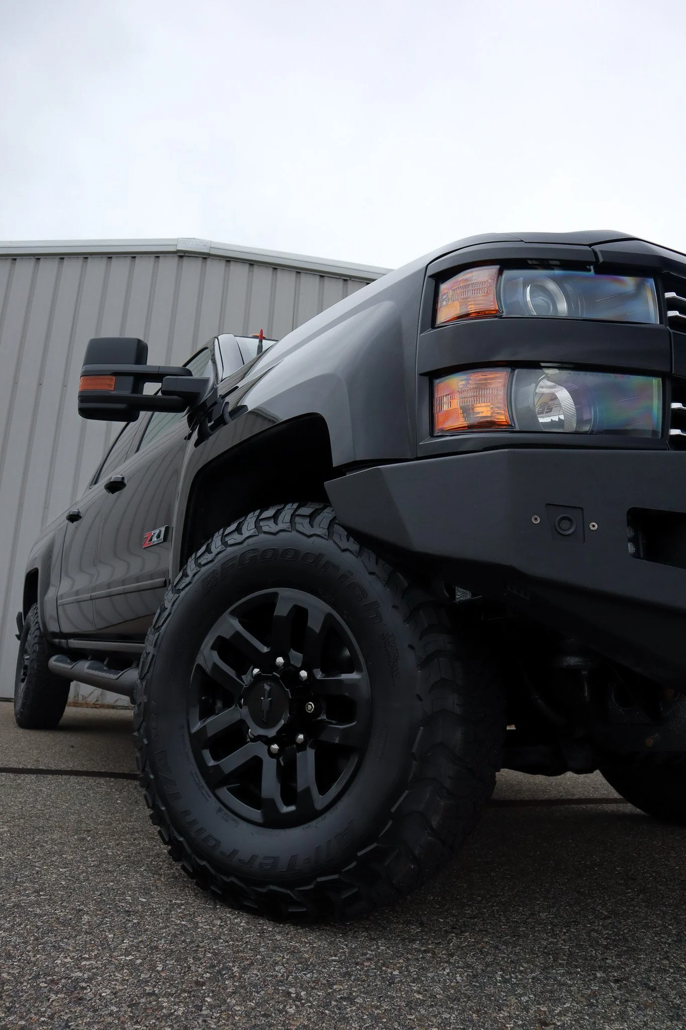 Close-up of a black pickup truck with large off-road tires, parked outdoors on a paved surface with a gray industrial building in background.