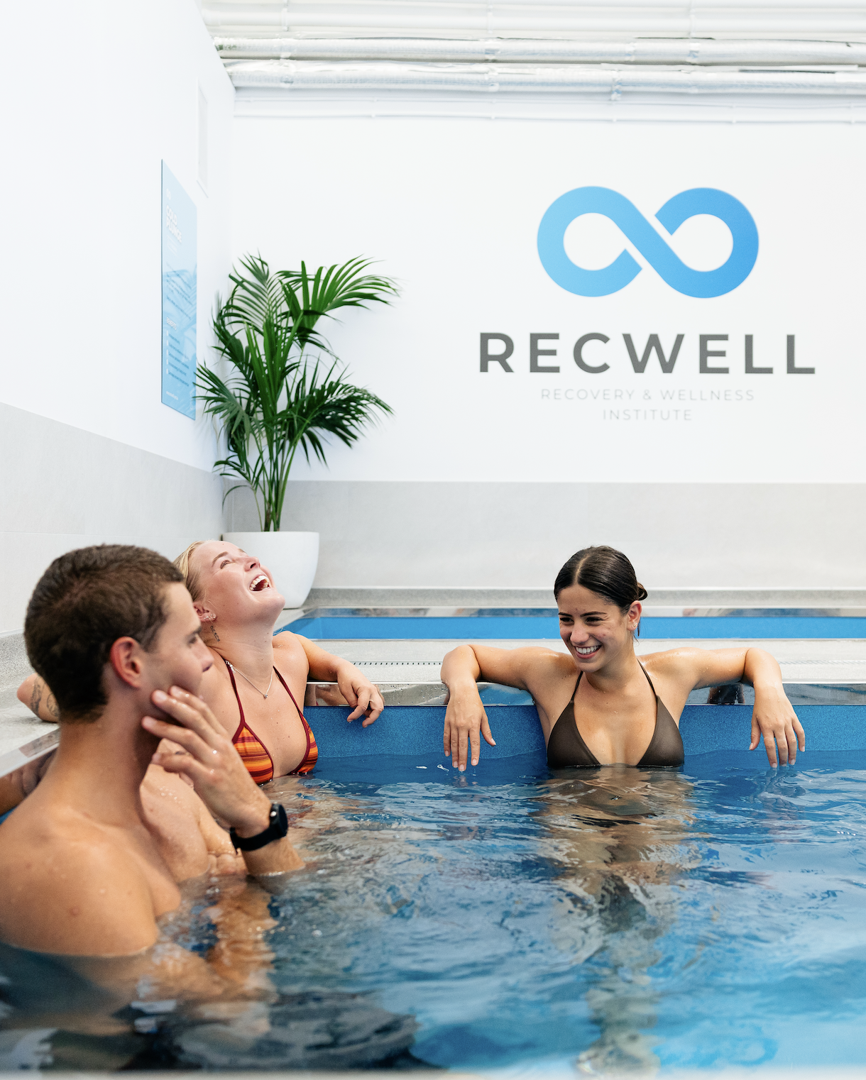 Three people laughing and enjoying themselves in an ice bath at Recwell Recovery & Wellness Institute, with a large green plant and a logo on the wall in the background.