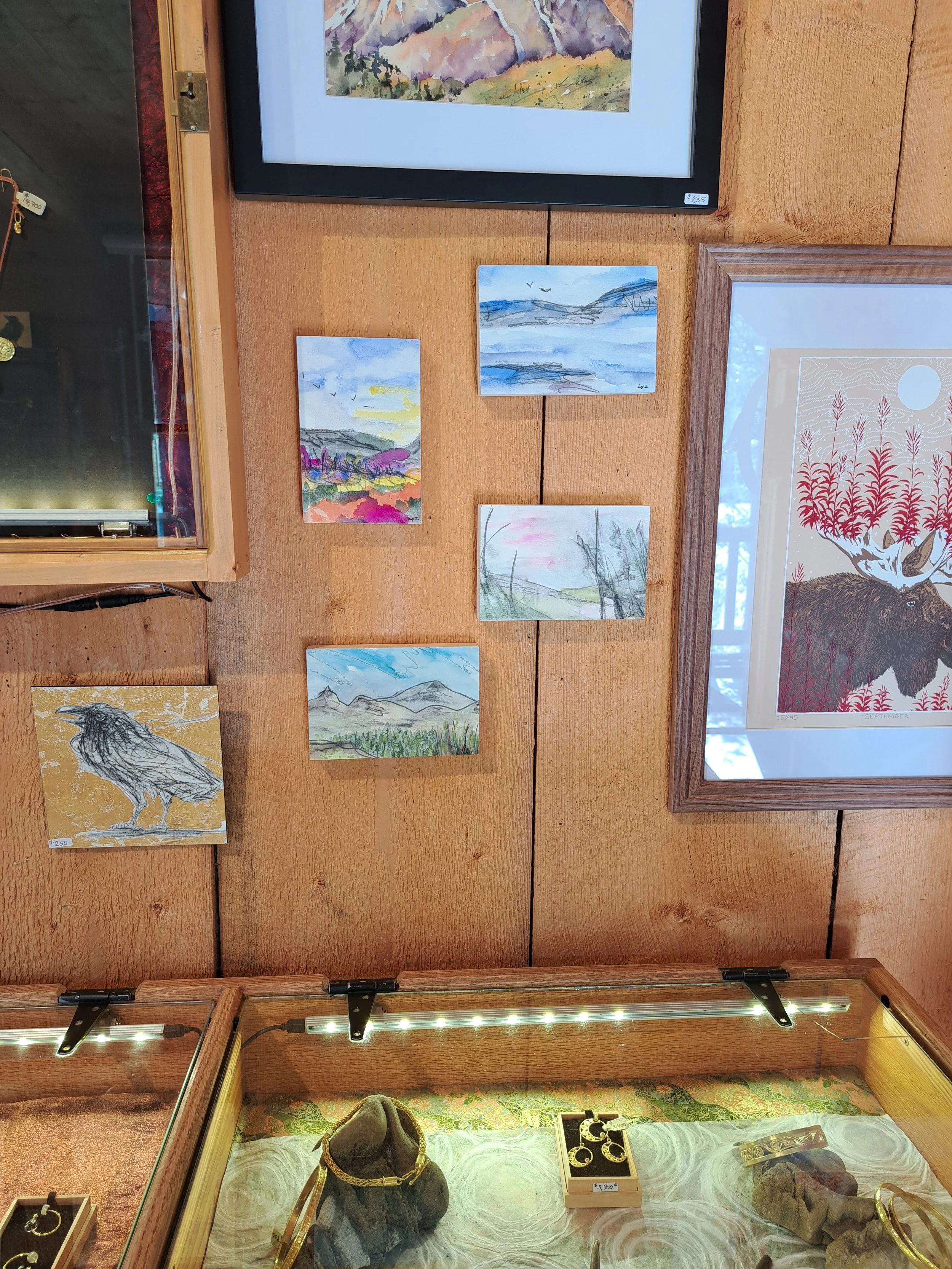 Display of five small landscape paintings of mountains, lakes, and colorful scenery on a wooden wall, with jewelry in a glass case below.
