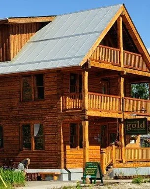 A two-story wooden cabin with a metal roof, wooden balconies, and a small porch, set against a clear blue sky.