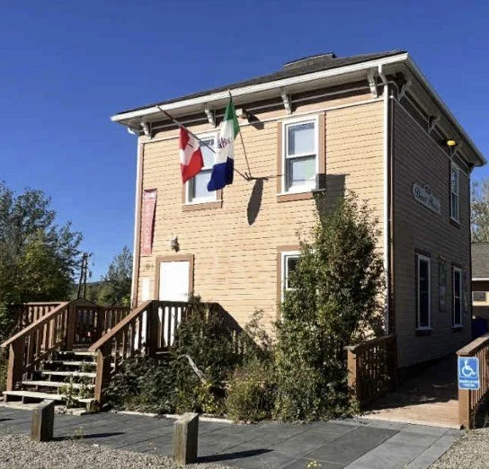 A beige two-story building with a small staircase leading to the front door, two flags hanging from the second floor, and a blue handicap parking sign nearby.