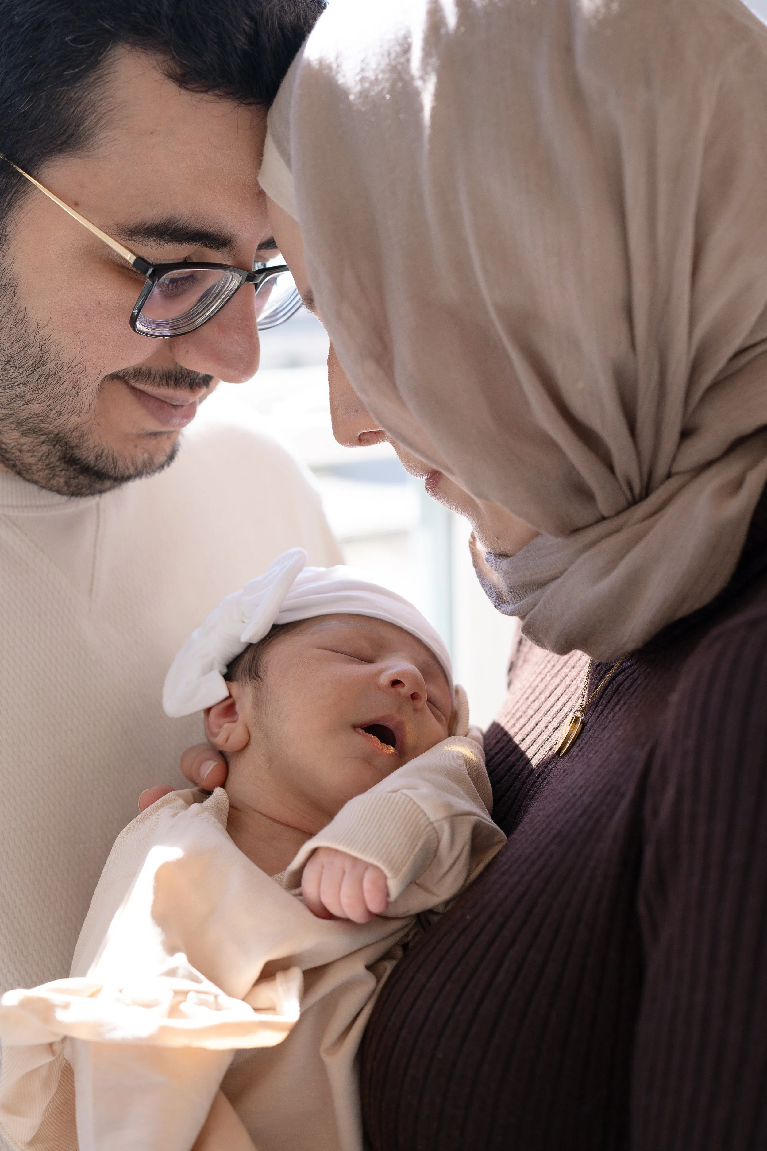 A man with glasses, a woman wearing a beige hijab, and a newborn baby in their arms, sharing a close, tender moment indoors.