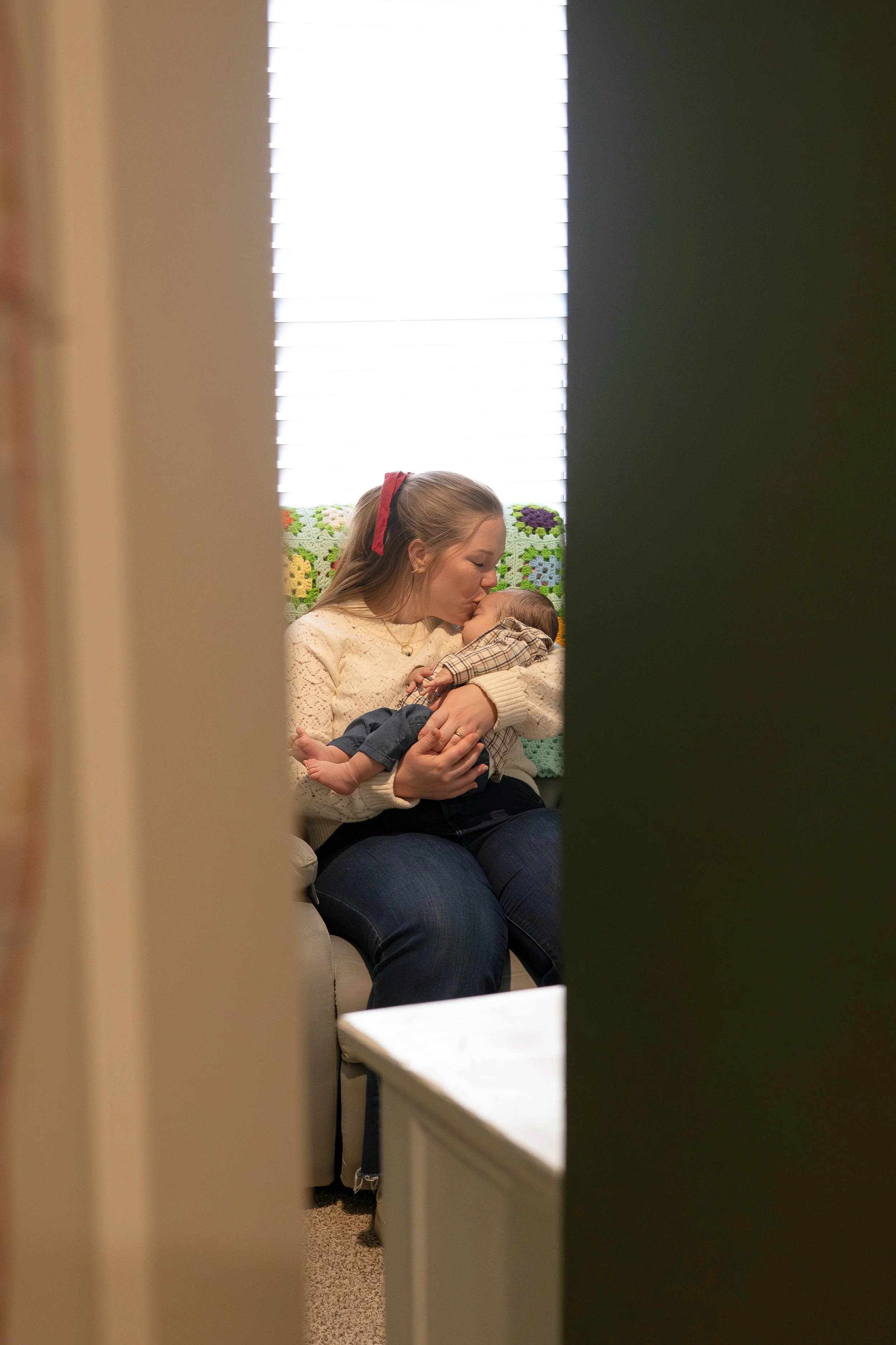 A woman sitting on a beige couch, holding a small child and kissing their forehead, seen through a partially opened door.
