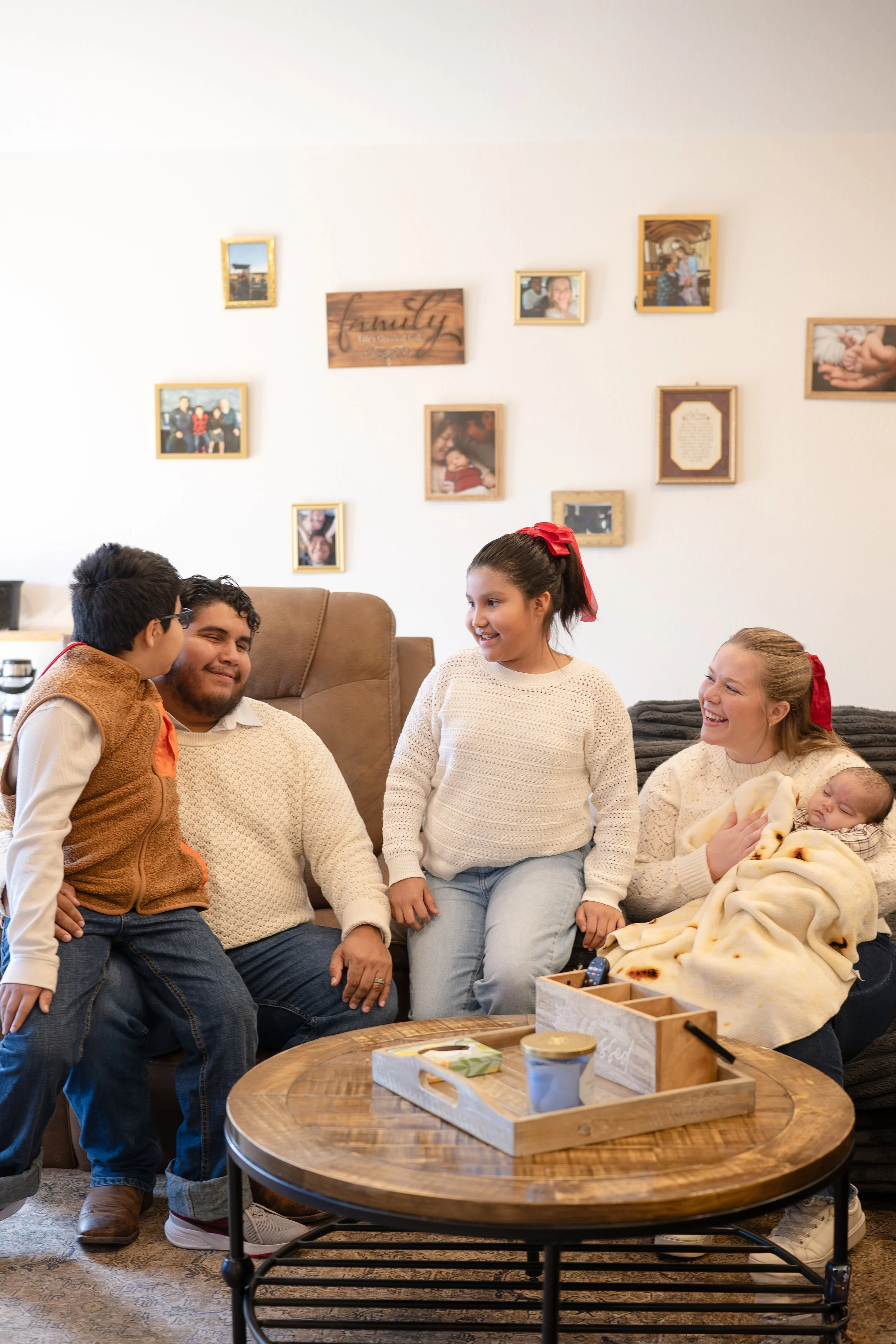 Family gathering in living room with children and baby, smiling and interacting, cozy setting with framed photos on wall, coffee table with snacks.