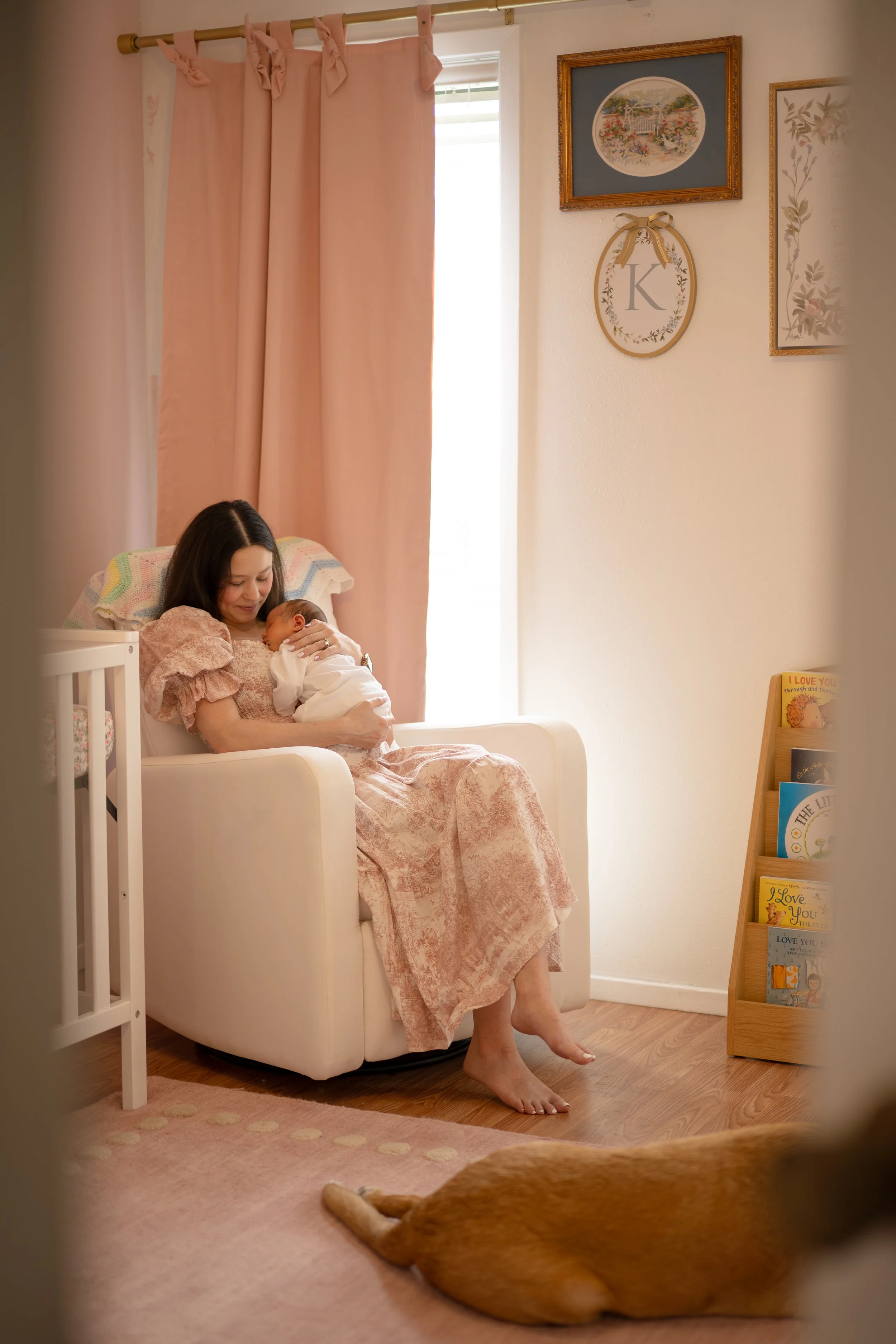 A woman sitting in a pink armchair, holding a newborn baby, in a cozy nursery with pink curtains, framed pictures on the wall, a bookshelf with children's books, and a dog lying on the floor.