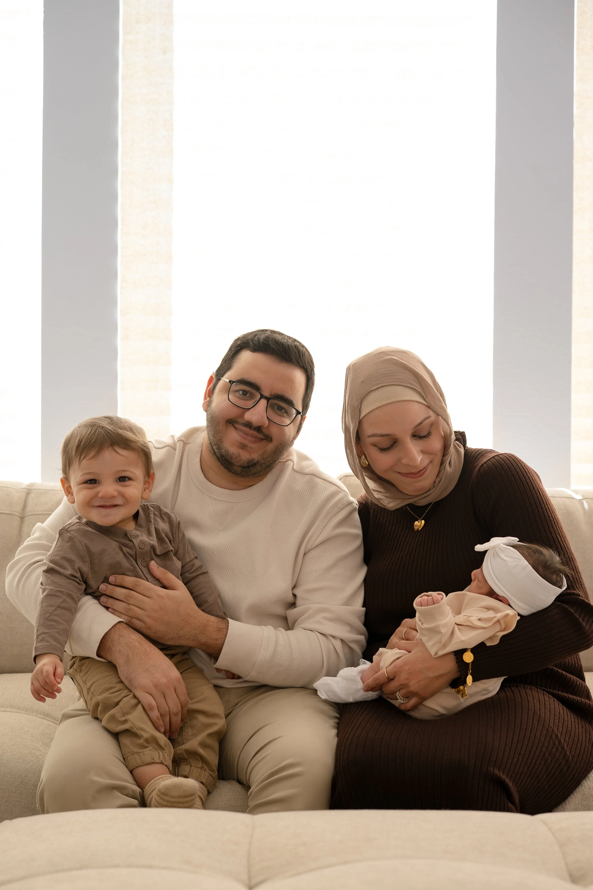 Family of four sitting on a sofa in a bright room, smiling at the camera. The father is holding a young boy, and the mother is holding a newborn girl.