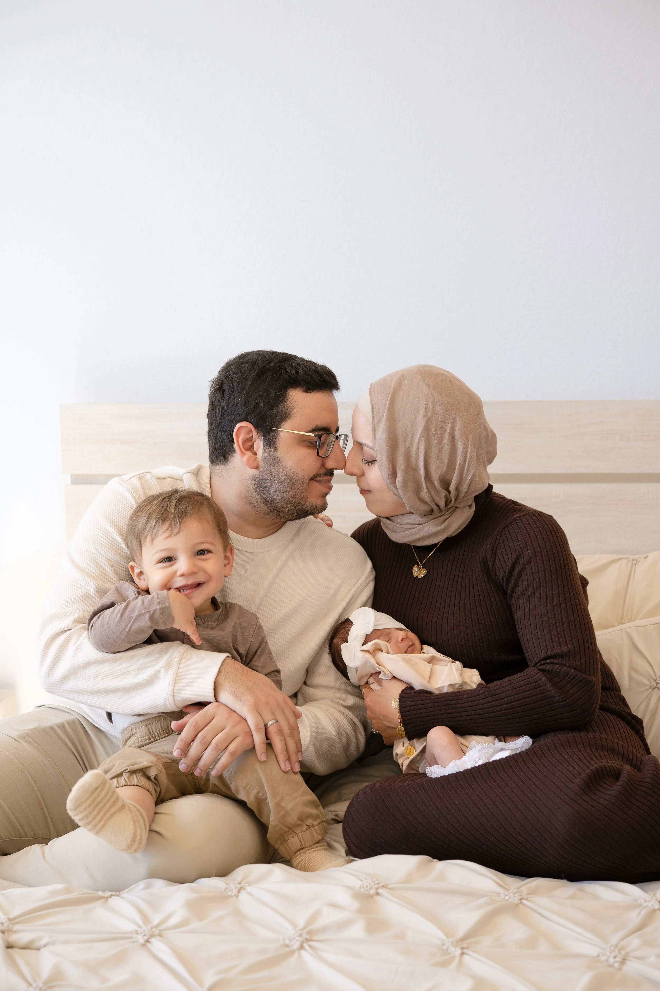 A family of four sitting on a bed, with a mother holding a newborn baby, a father leaning close, and a toddler smiling at the camera.
