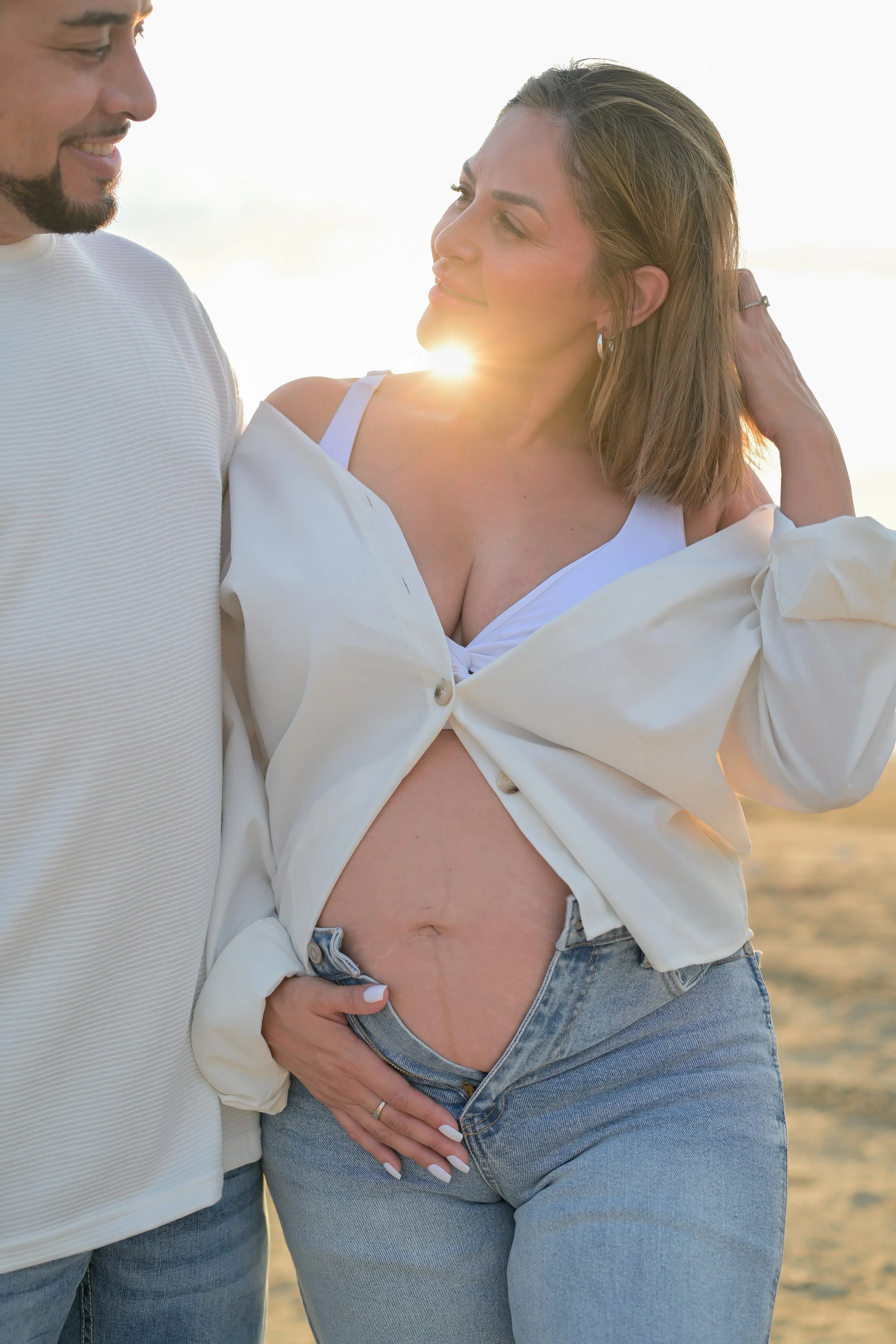 A woman with visible stretch marks on her abdomen, wearing a white tank top and jeans, standing on a sandy beach at sunset with a man. The man is smiling and holding her waist, and she has her hand on his chest while looking at him.
