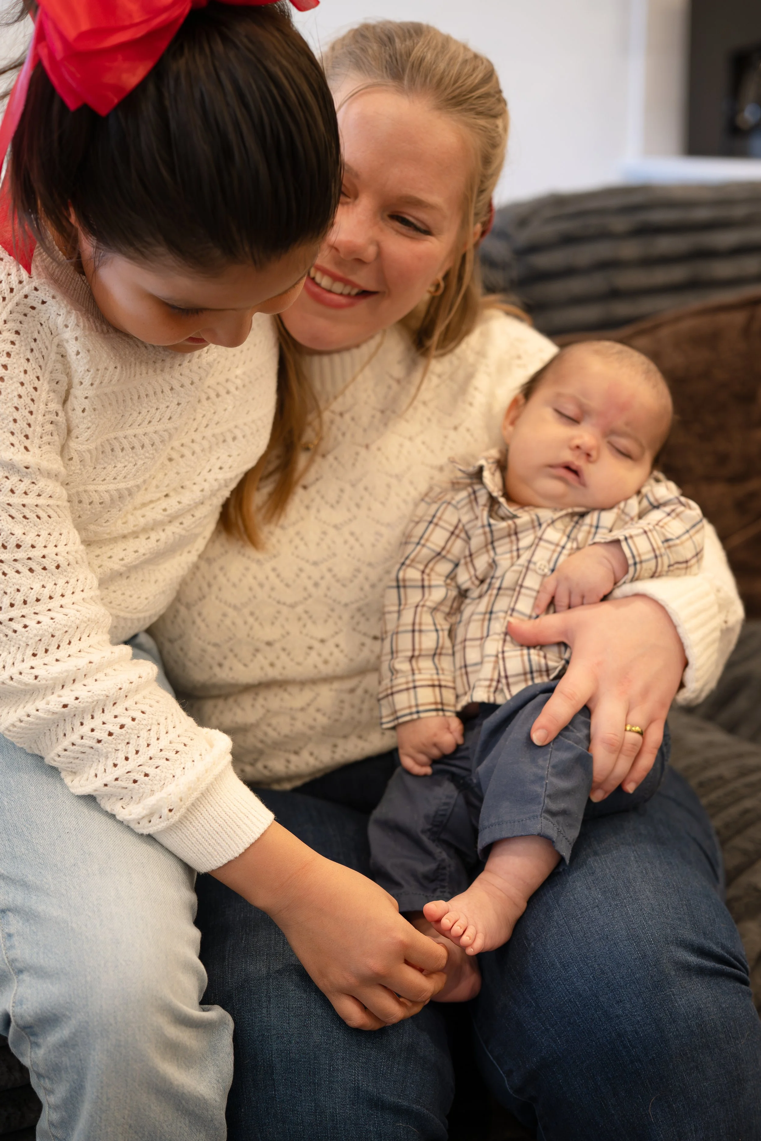 A woman with long red hair holding a sleeping baby dressed in a plaid shirt and navy pants, while a girl in a white crochet sweater leans over and interacts with the baby, all sitting on a dark-colored couch.