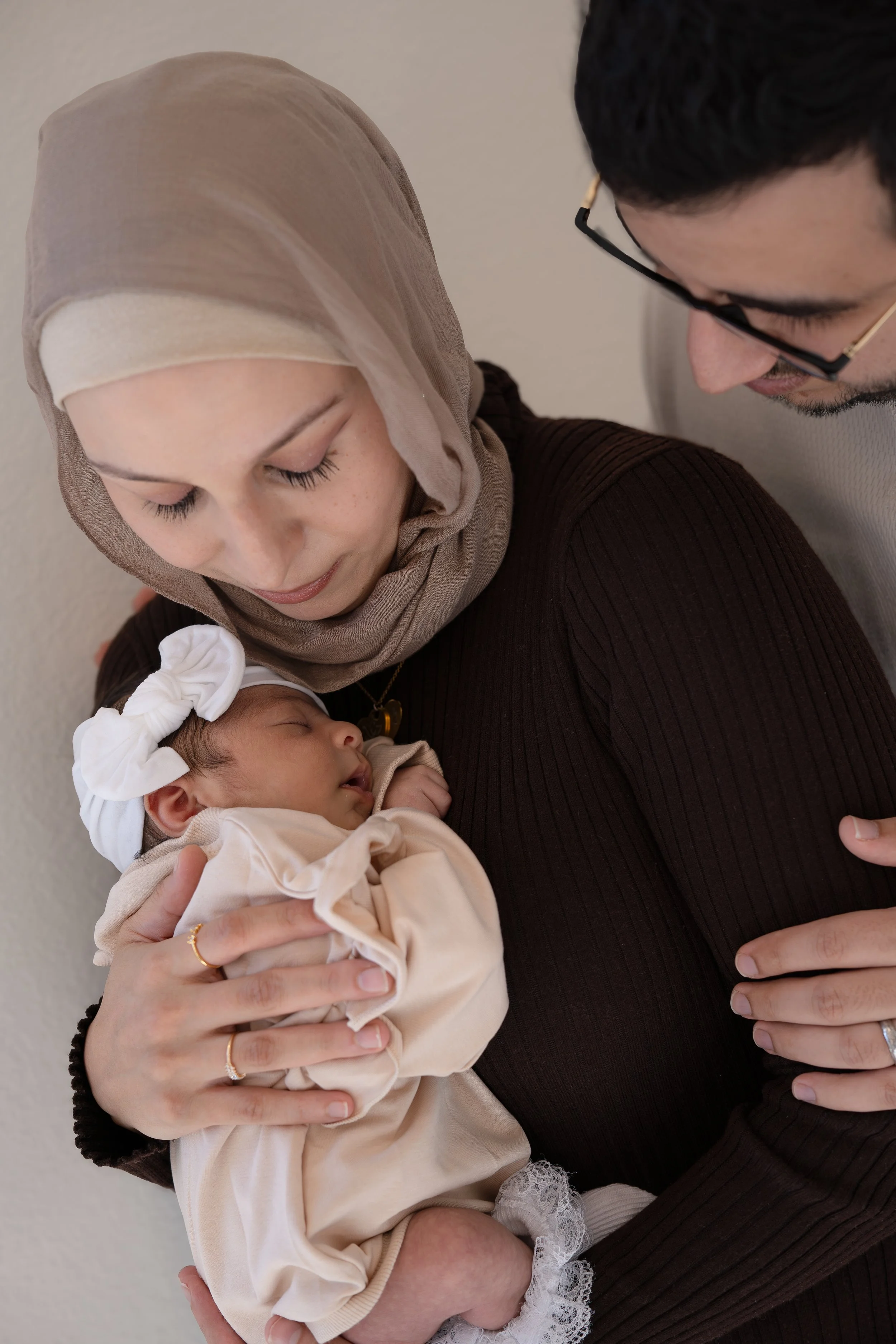 A woman wearing a beige hijab holding a sleeping baby dressed in beige with a large white bow on her head, with a man leaning over and smiling.
