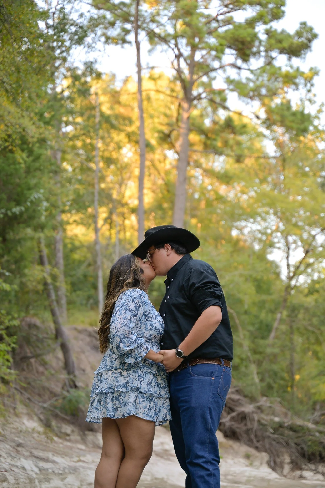 A couple kissing outdoors in a wooded area, with greenery and trees in the background.