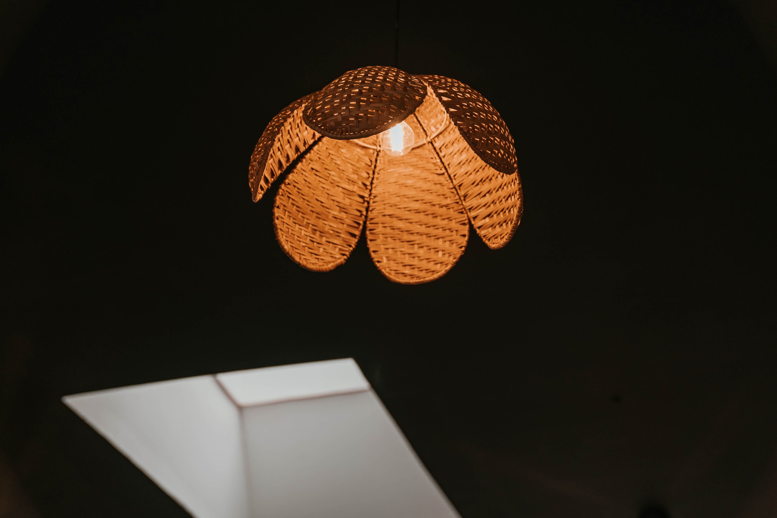 A woven pendant light hanging from the ceiling with a visible light bulb inside, illuminating the surrounding dark space.