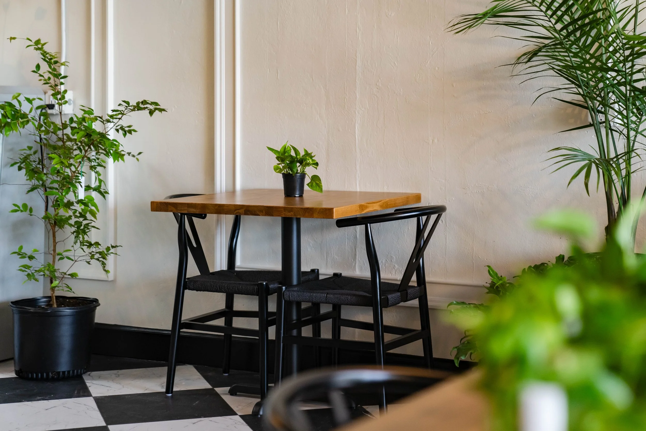 A small wooden table with a potted plant on top, surrounded by larger potted plants, in a room with a black and white checkered floor and a plain beige wall.