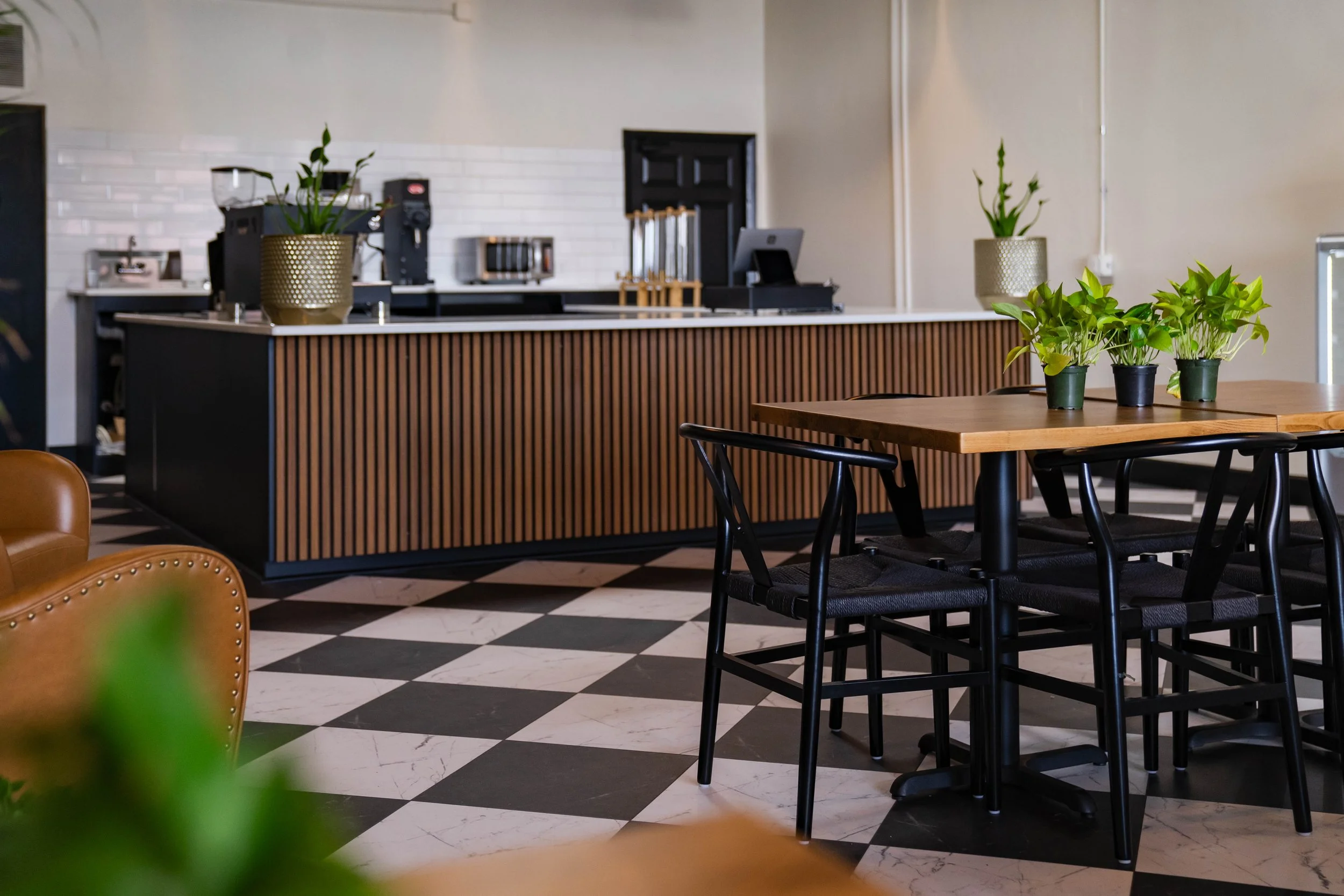 A stylish cafe interior featuring a checkered black and white floor, a wooden top table with black chairs, potted plants on the table and counter, and a coffee machine behind the counter with a white brick wall.