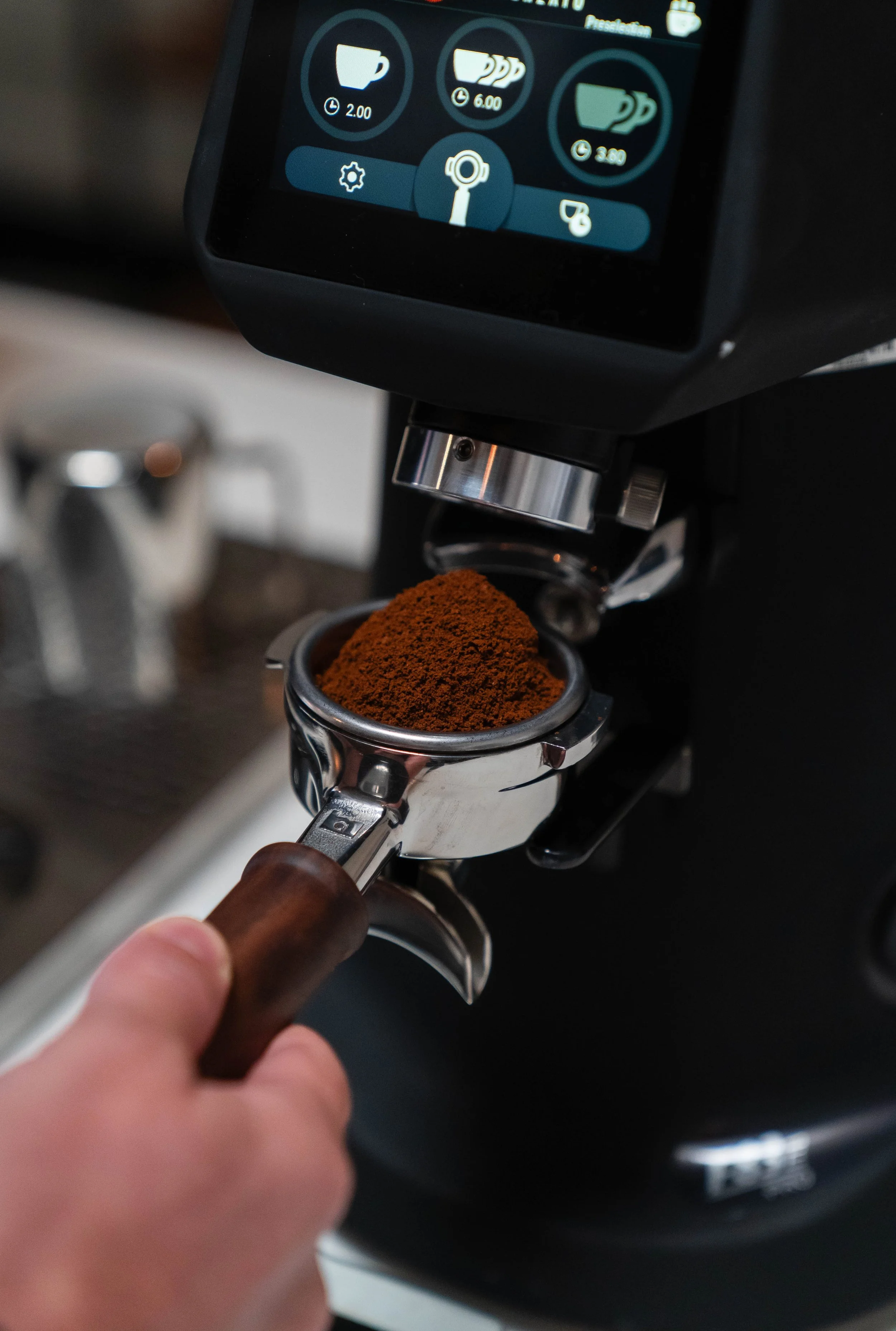 Close-up of a hand holding a portafilter with freshly ground coffee underneath a commercial espresso machine's grinder. The machine's digital display shows coffee options and prices.