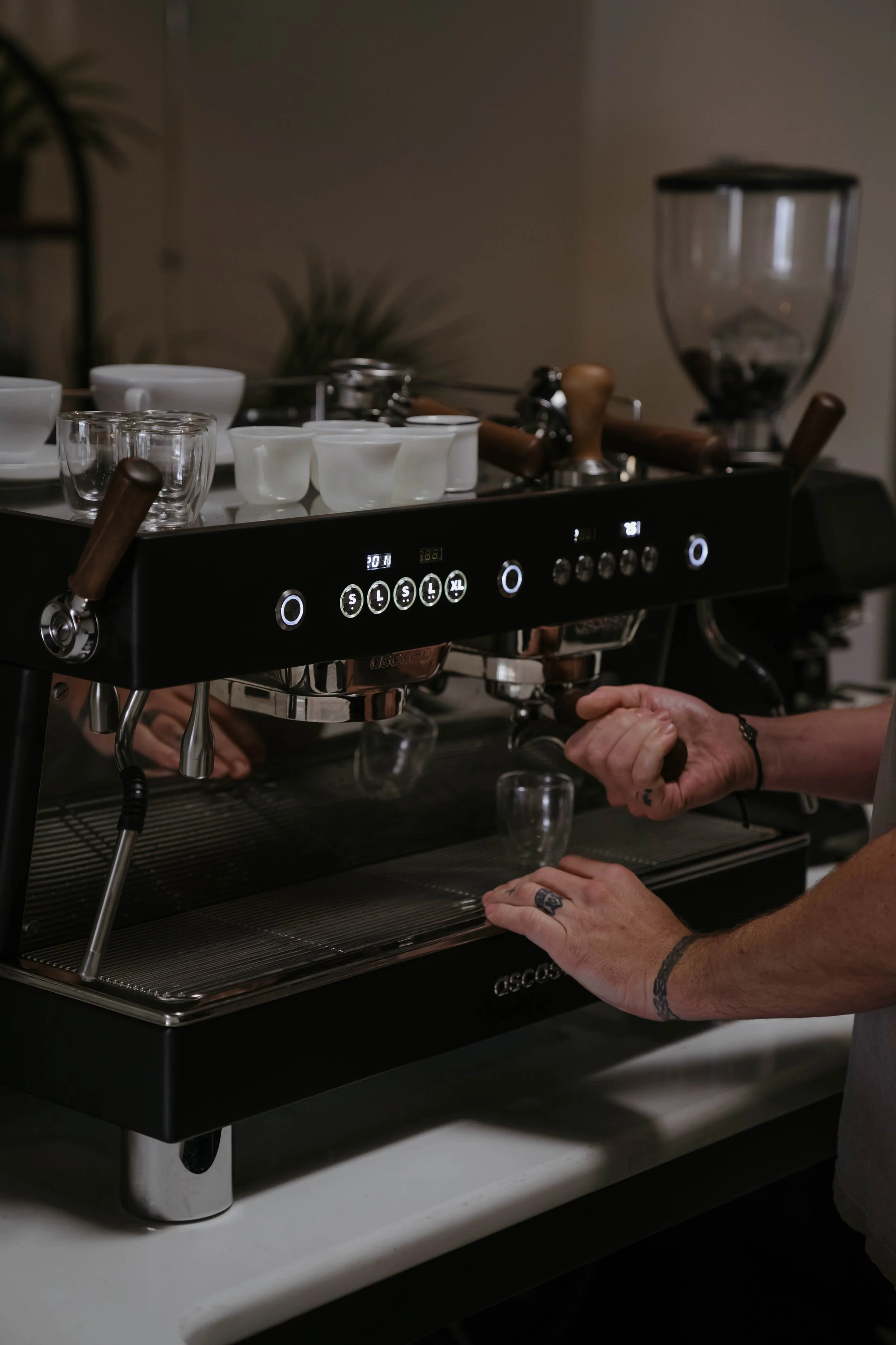 A person operating a professional espresso machine, with hands on the machine, pouring or preparing coffee cups, in an indoor setting.