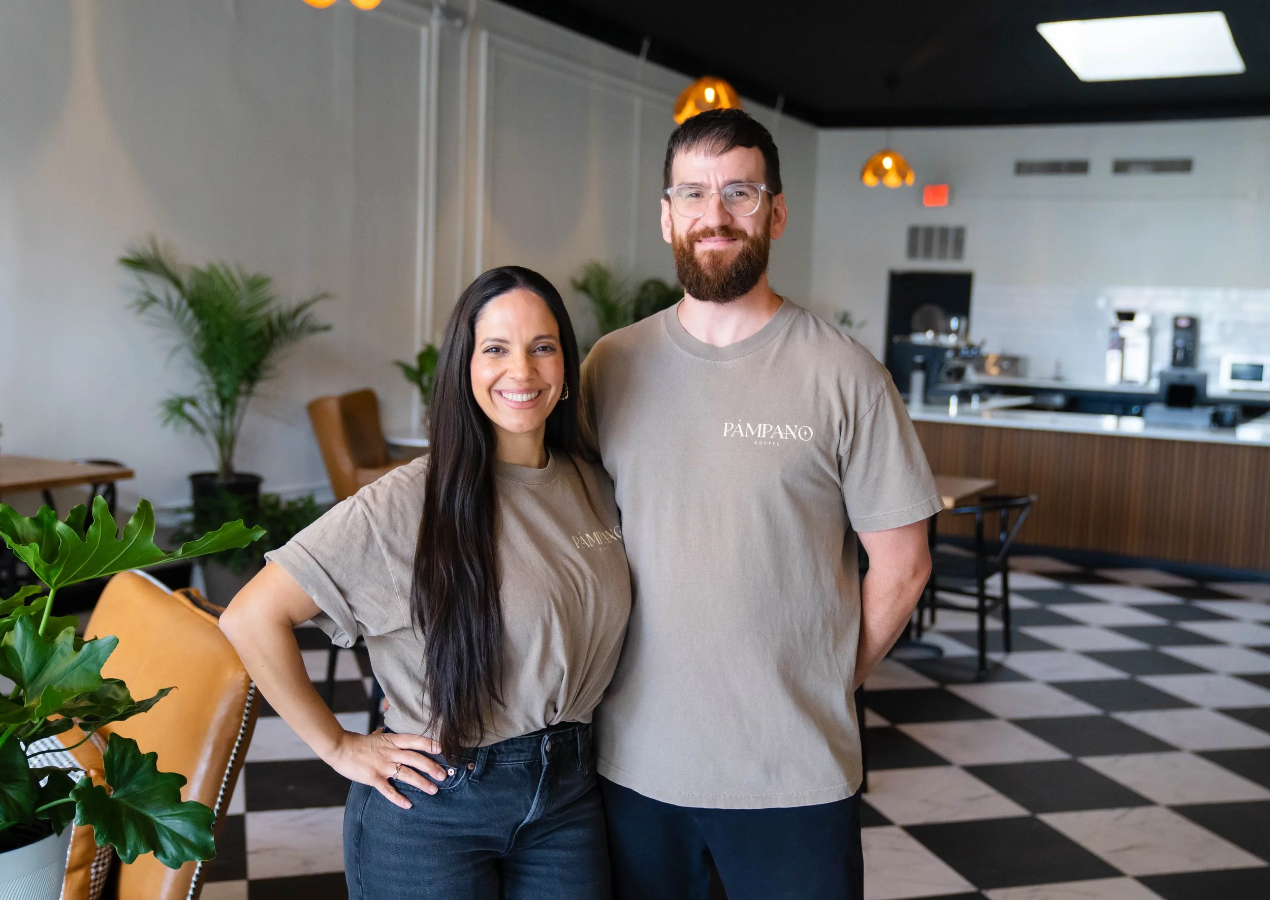 Two people standing inside a coffee shop, smiling at the camera. The woman on the left has long dark hair and is wearing a beige shirt and jeans. The man on the right has a beard, glasses, and short dark hair, wearing a beige t-shirt with 'PÁMPANO' written on it. The background features a modern cafe interior with black-and-white checkered flooring, potted plants, and warm lighting.