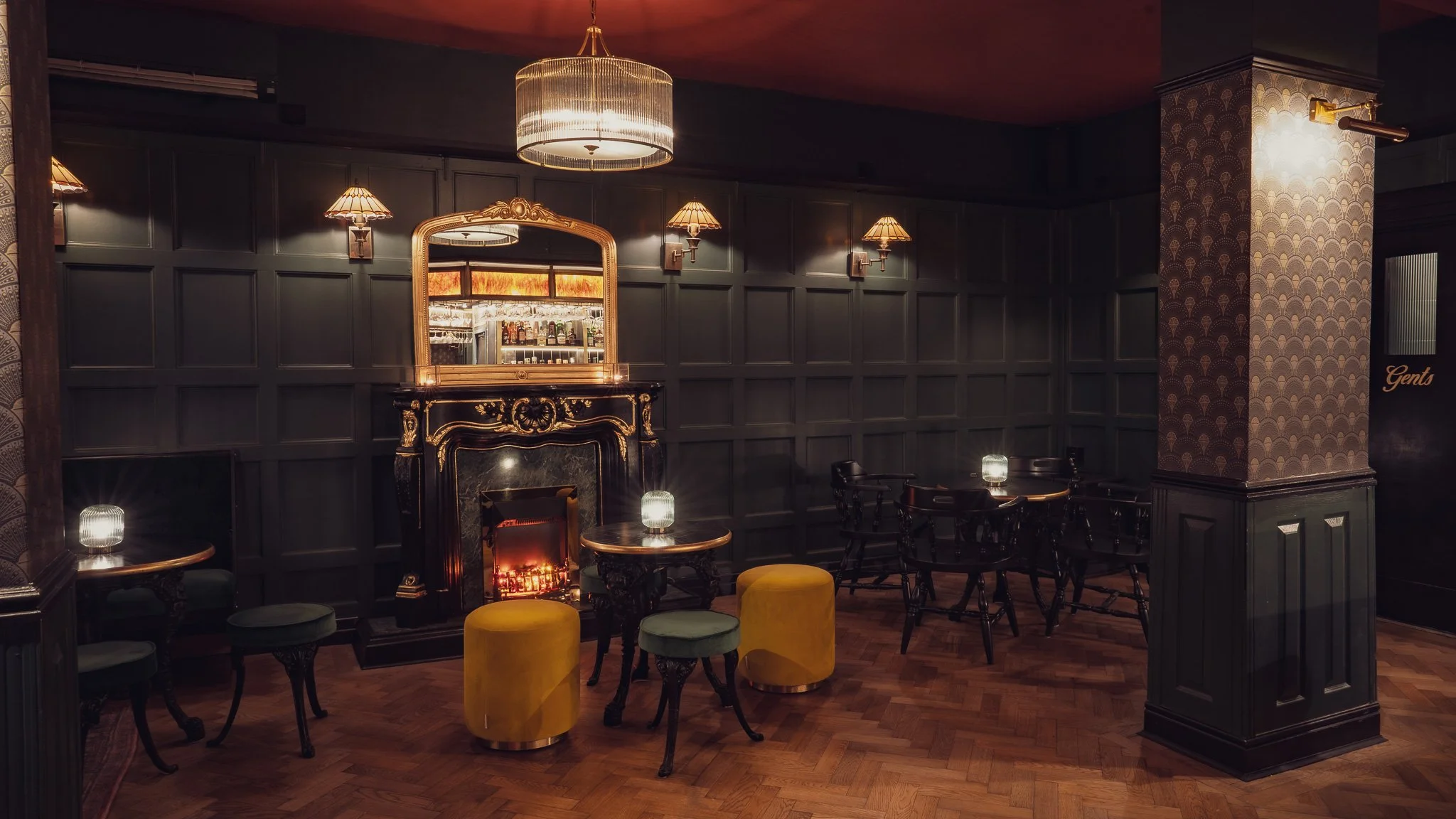 Elegant bar lounge with dark paneled walls, a fireplace, small tables with lamps, and upholstered stools, creating a cozy, vintage atmosphere.