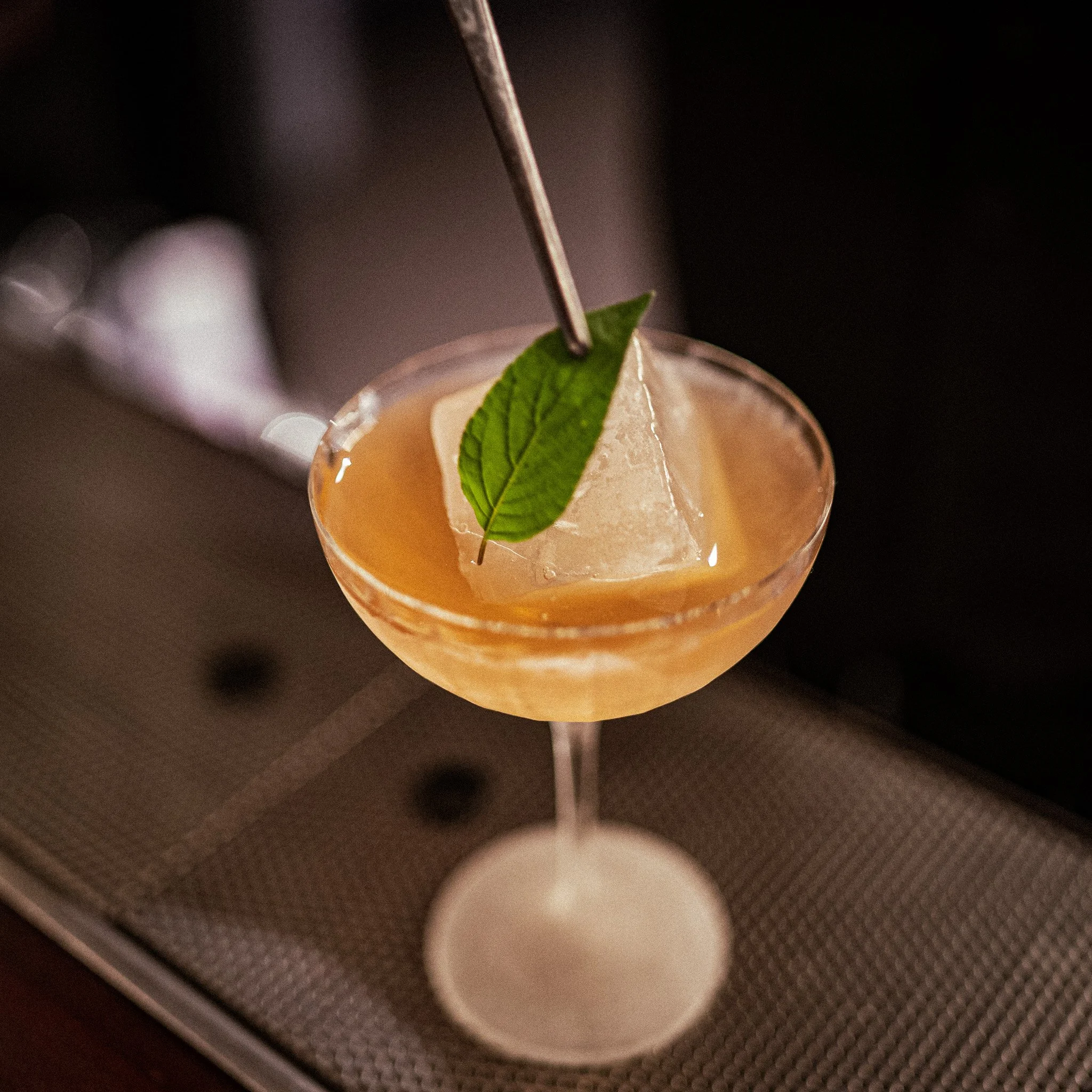 A cocktail in a coupe glass with a large ice cube, garnished with a green mint leaf and a metal stirrer.