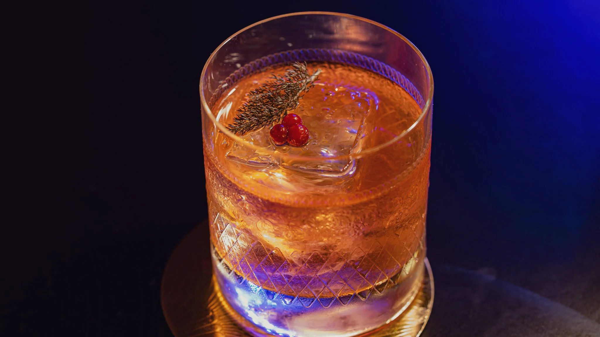 A glass of orange-colored cocktail garnished with a sprig of herbs, red berries, and ice cubes, on a dark surface with blue lighting.