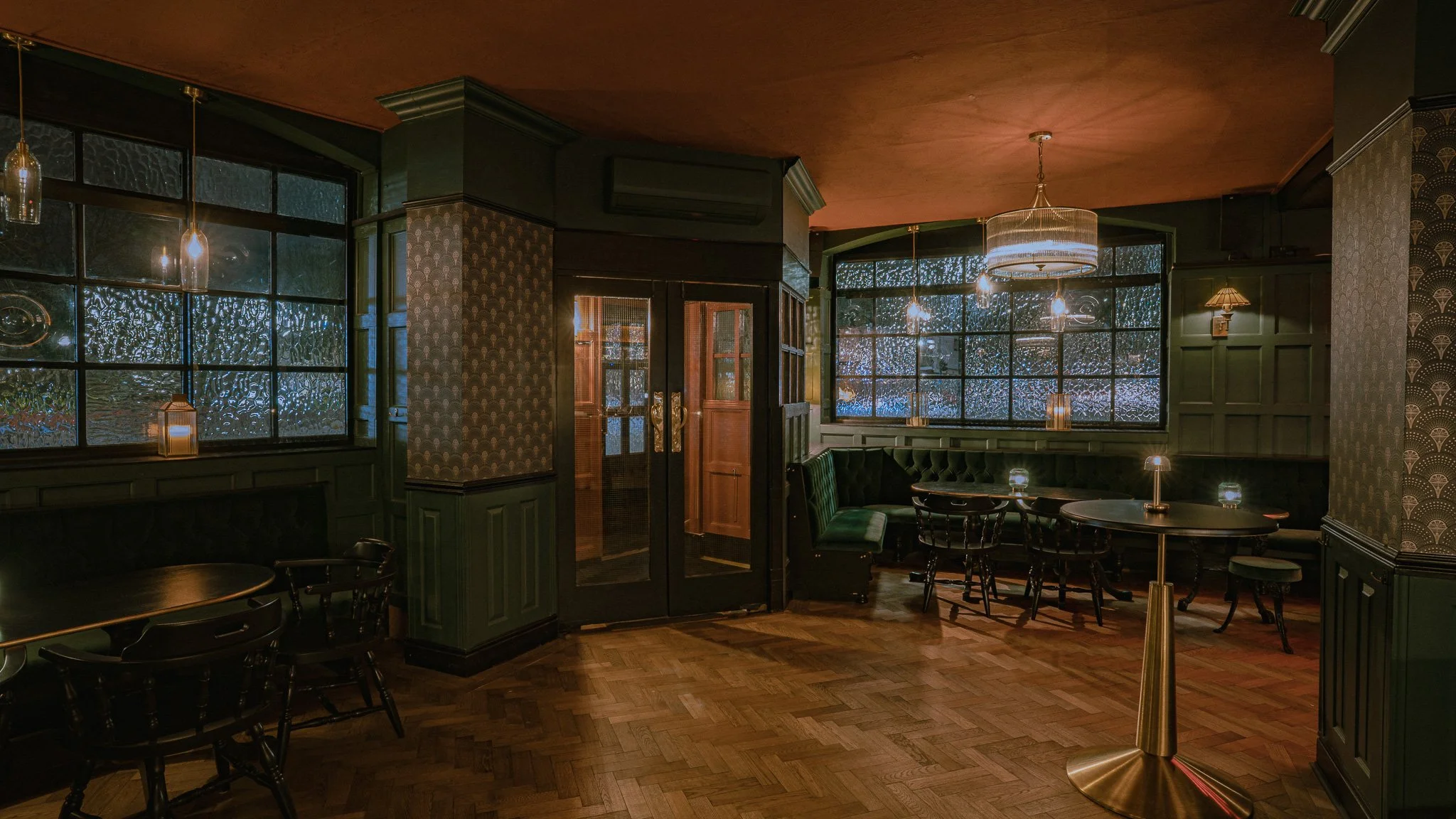 Dimly lit interior of a cozy restaurant or bar with dark green walls, patterned wallpaper, and warm wooden floors. There are tables with chairs and decorative hanging lights, some candles on the windowsills, and a large glass door.
