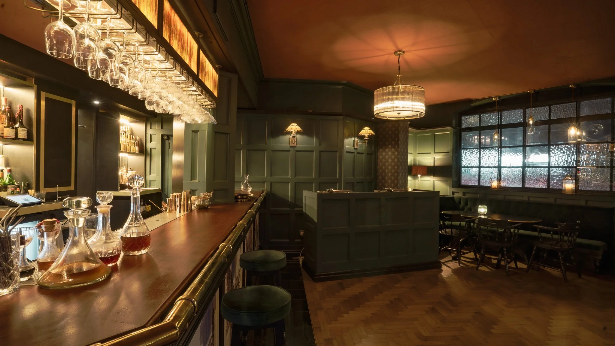 An empty bar with dark green walls, brass accents, a wooden countertop, and hanging glassware, in a warmly lit room with wood flooring and large textured windows.