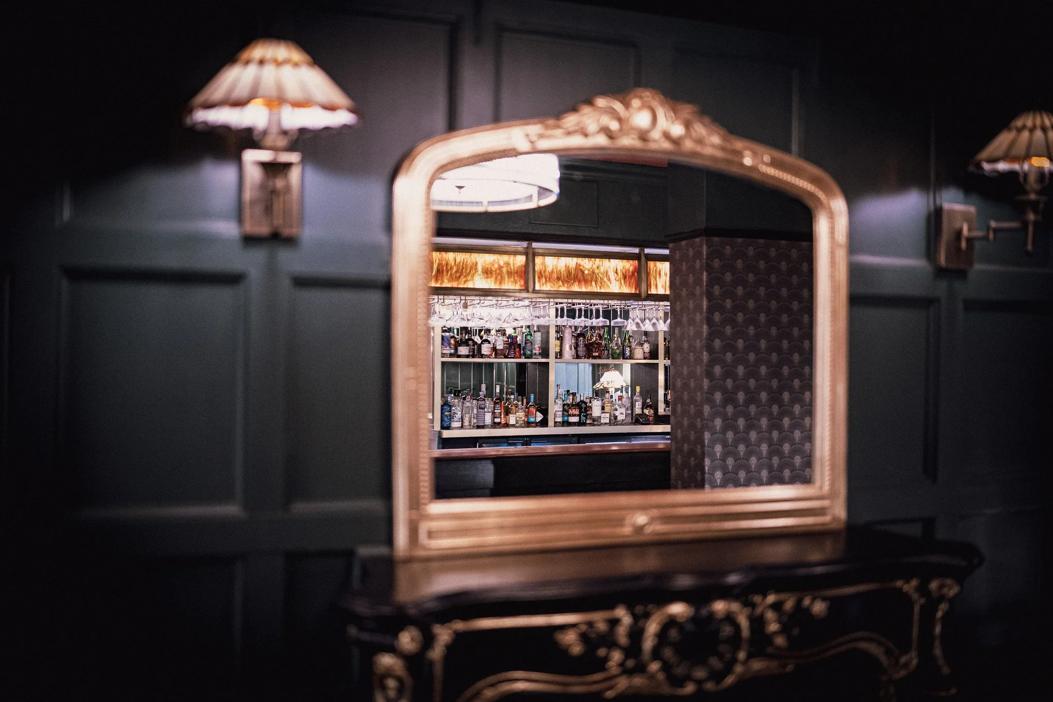 Bar scene reflected in a decorative mirror with dark walls and warm lighting.