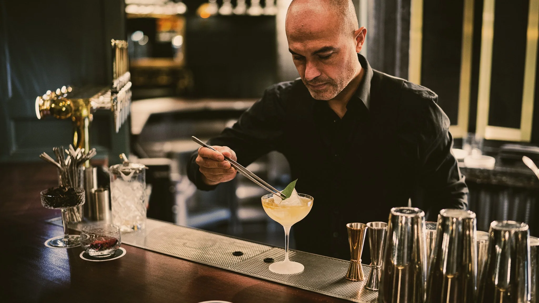 A bartender in a black shirt preparing a cocktail at a bar with bar tools and glasses.