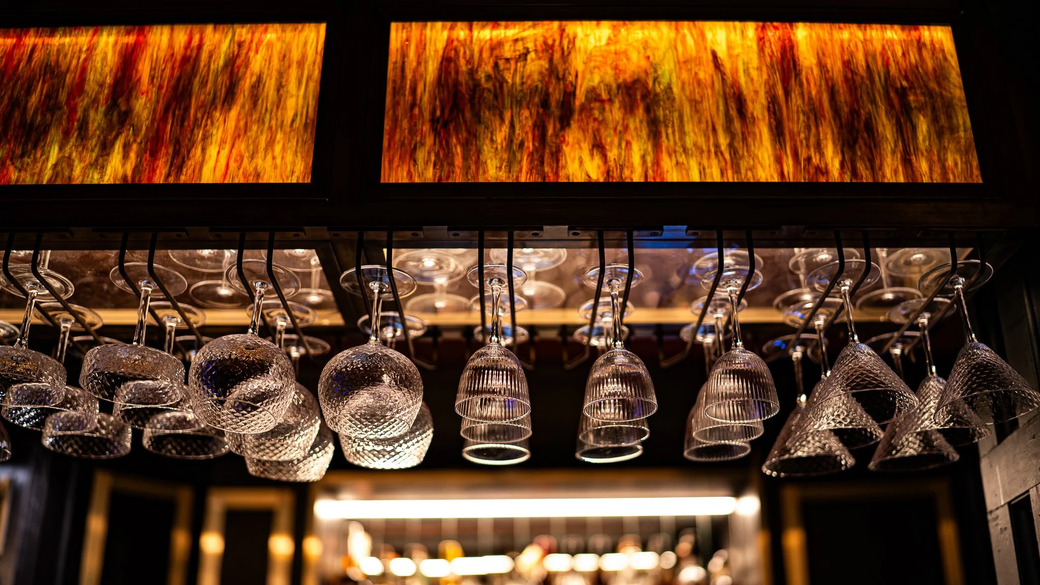 Hanging glassware including wine glasses and tumblers under a wooden rack in a bar or restaurant setting.